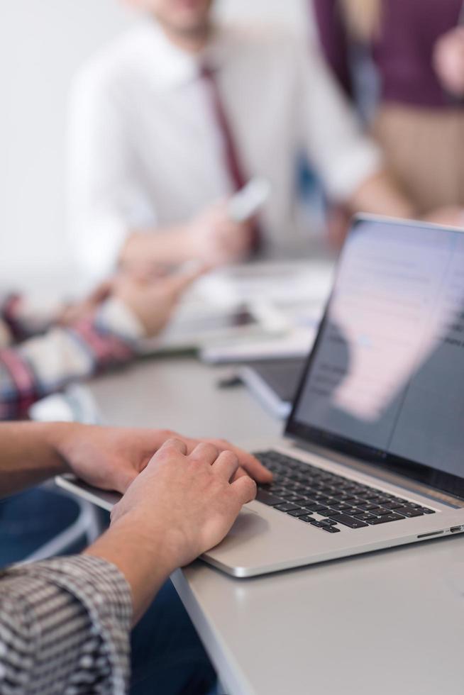 close up of business man hands typing on laptop with team on meeting in background photo