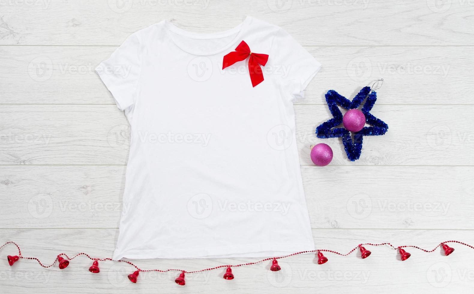 Close up white blank template t shirt with copy space and Christmas Holiday concept. Top view mockup t-shirt and red bow on white wooden background. Happy New Year decorations accessories. Xmas outfit photo