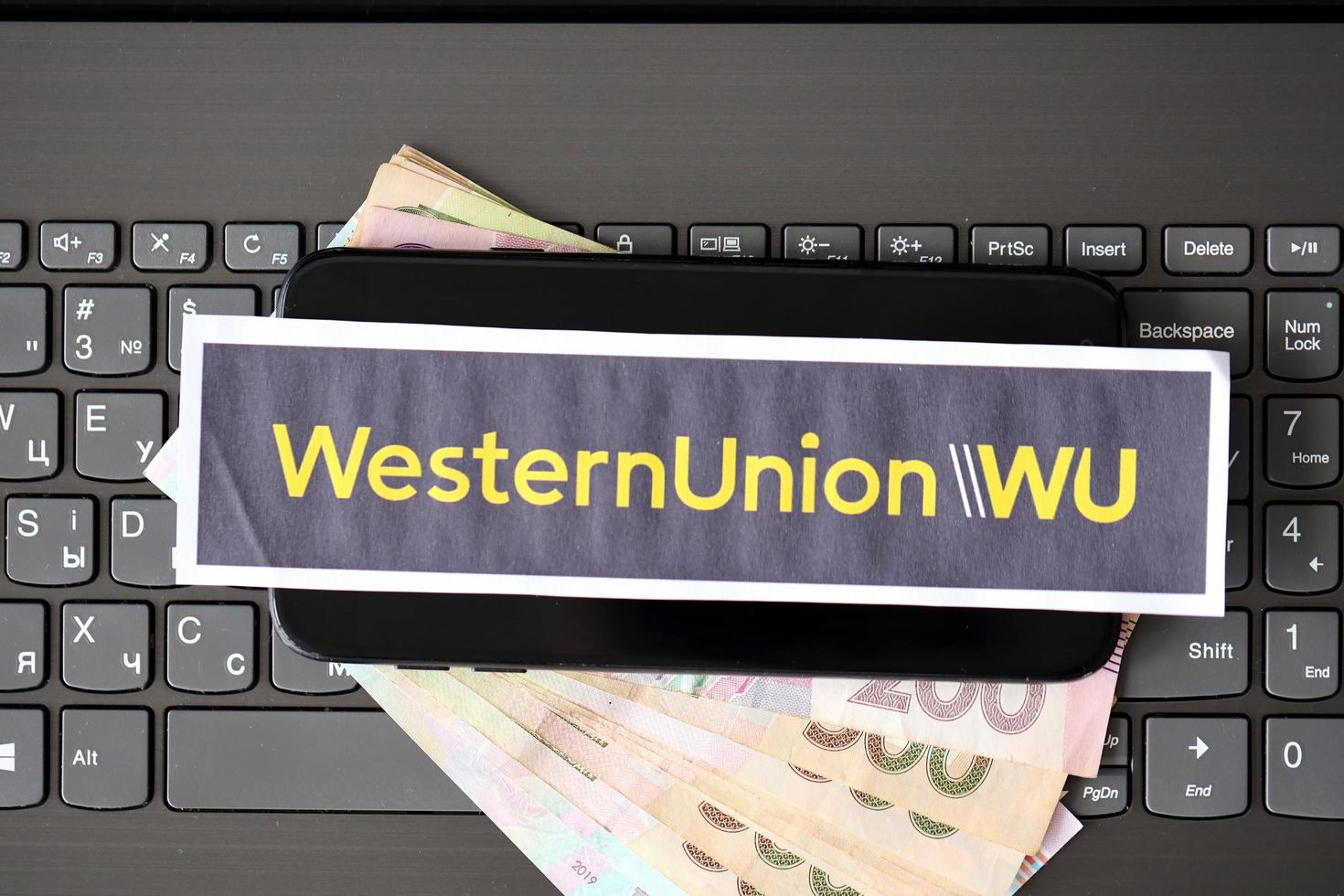 TERNOPIL, UKRAINE - SEPTEMBER 6, 2022 Western Union paper logotype lies on black laptop with ukrainian hryvnias. Western Union Company is American multinational financial services company photo