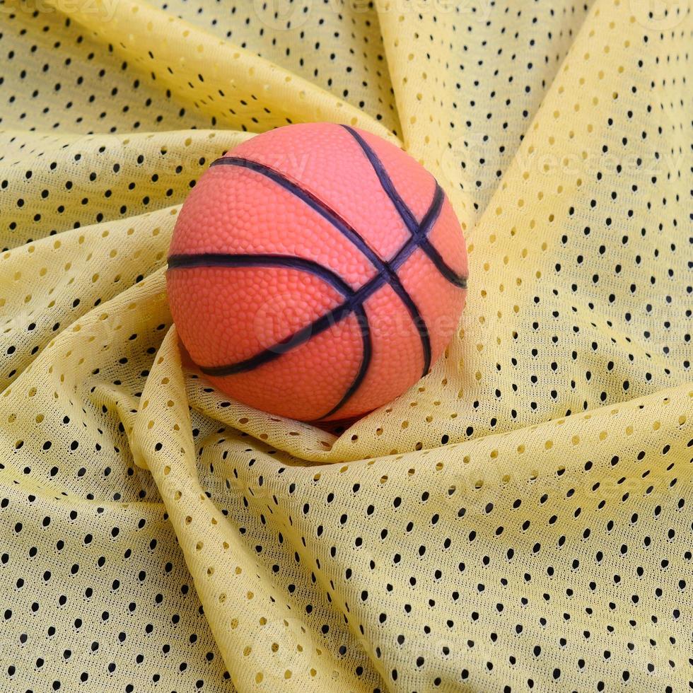 Small orange rubber basketball lies on a yellow sport jersey clothing fabric texture and background with many folds photo