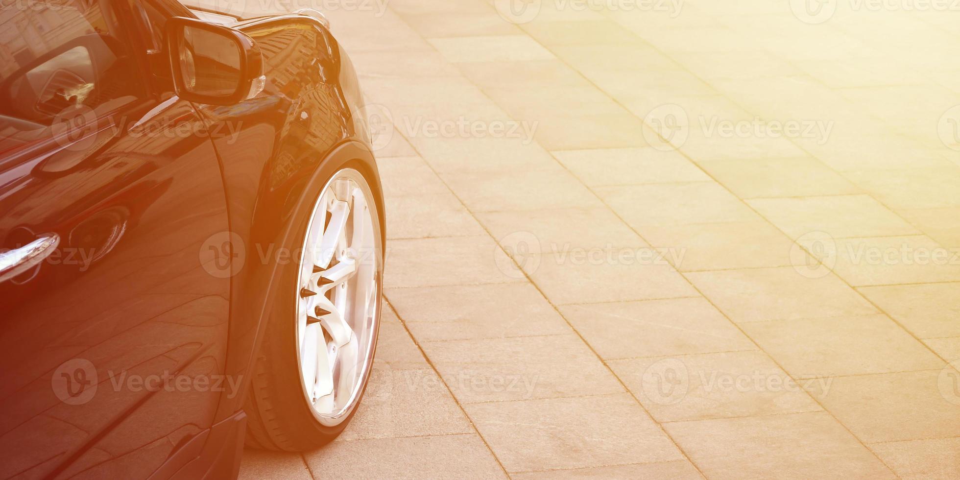 Diagonal view of a black glossy car with white wheels, which stands on a square of gray tiles photo