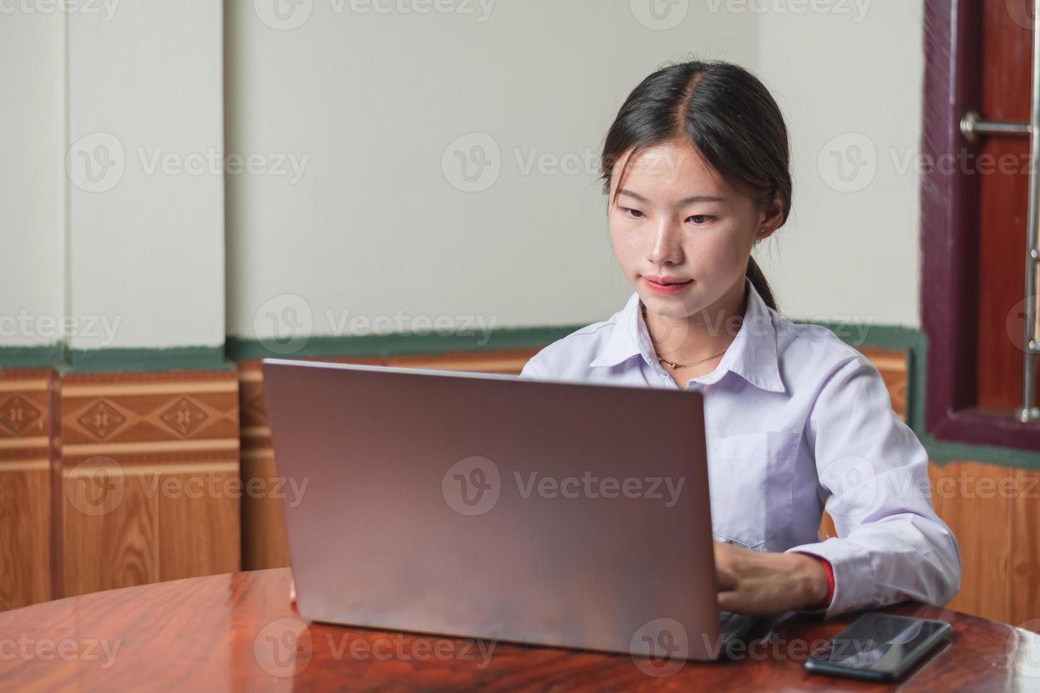 una estudiante con traje de estudiante de camisa blanca está escribiendo y aprendiendo en línea desde una computadora portátil en la casa, usando una computadora portátil e Internet inalámbrico en el escritorio. concepto de educación y estudio, toma de espacio de copia foto