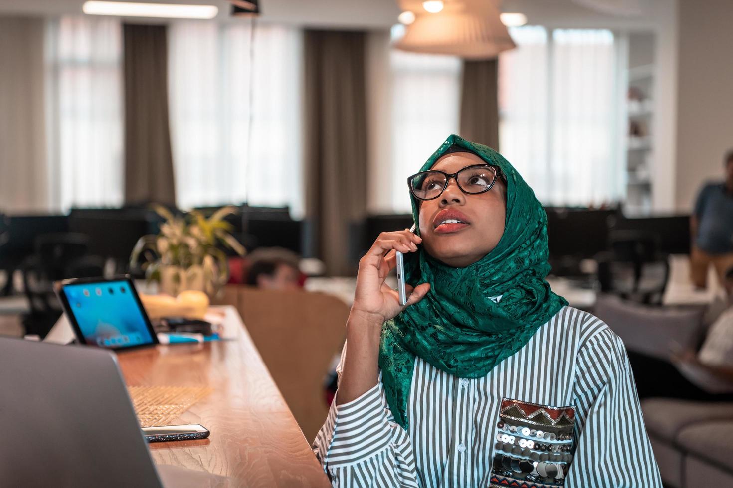 Smiling young woman talk on smartphone work on laptop look in distance thinking. Happy African American customer client use modern gadgets cellphone and computer at home office. photo