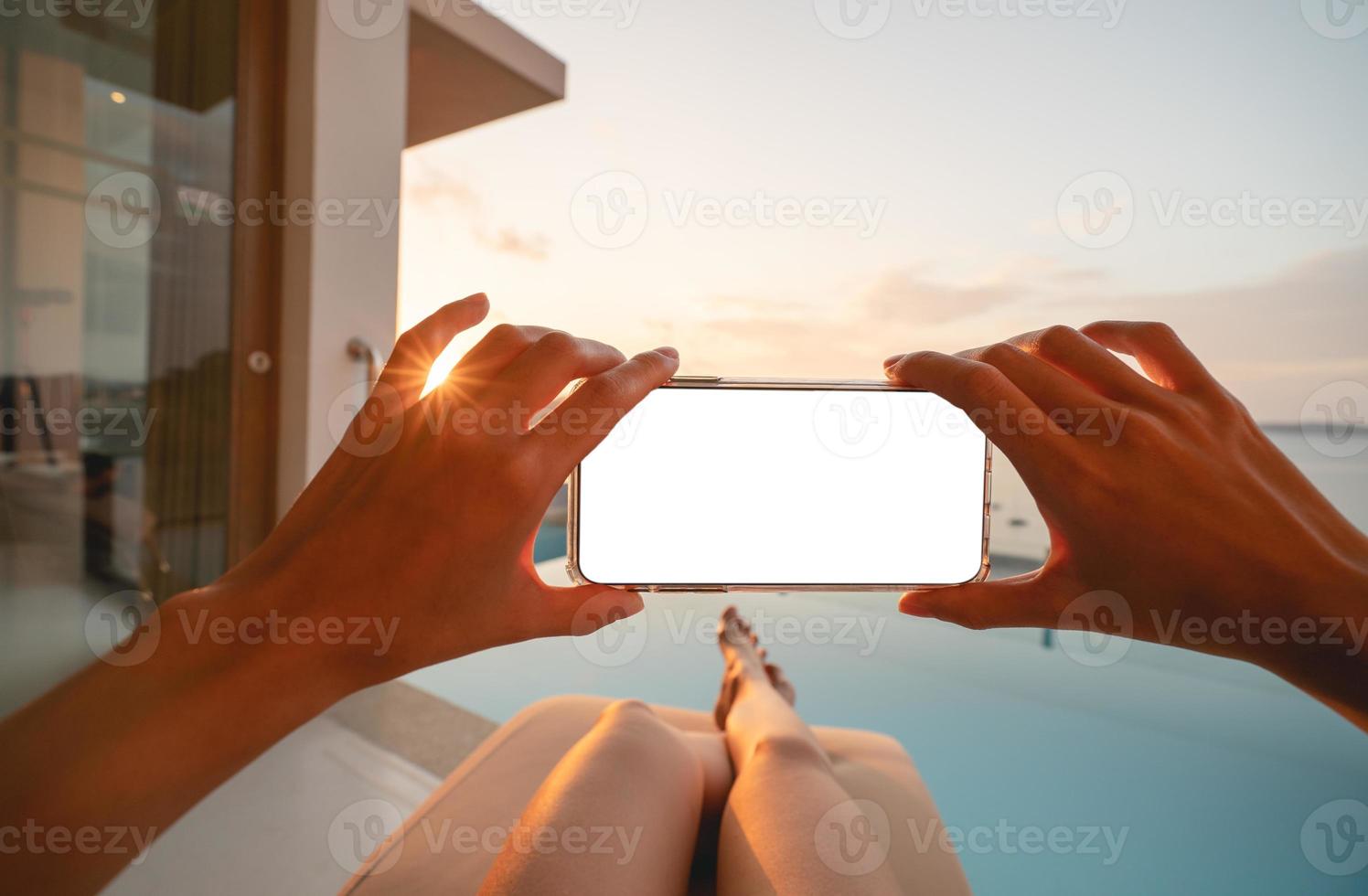 Woman holding mobile phone with blank screen while laying on sunbathing bed with beach view. photo
