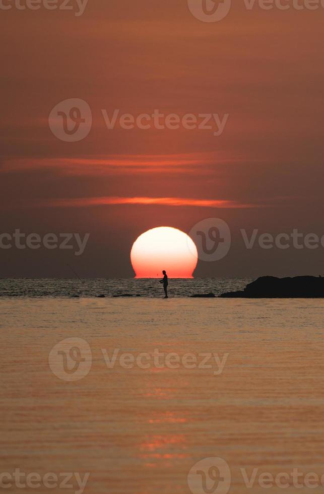 Sunset ocean view. Sun above the sea on orange sky. Fisherman with fishing rod on the rock. photo