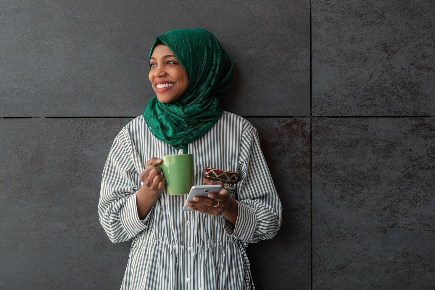 mujer de negocios musulmana africana con hiyab verde usando teléfono móvil durante el descanso para tomar café del trabajo afuera foto