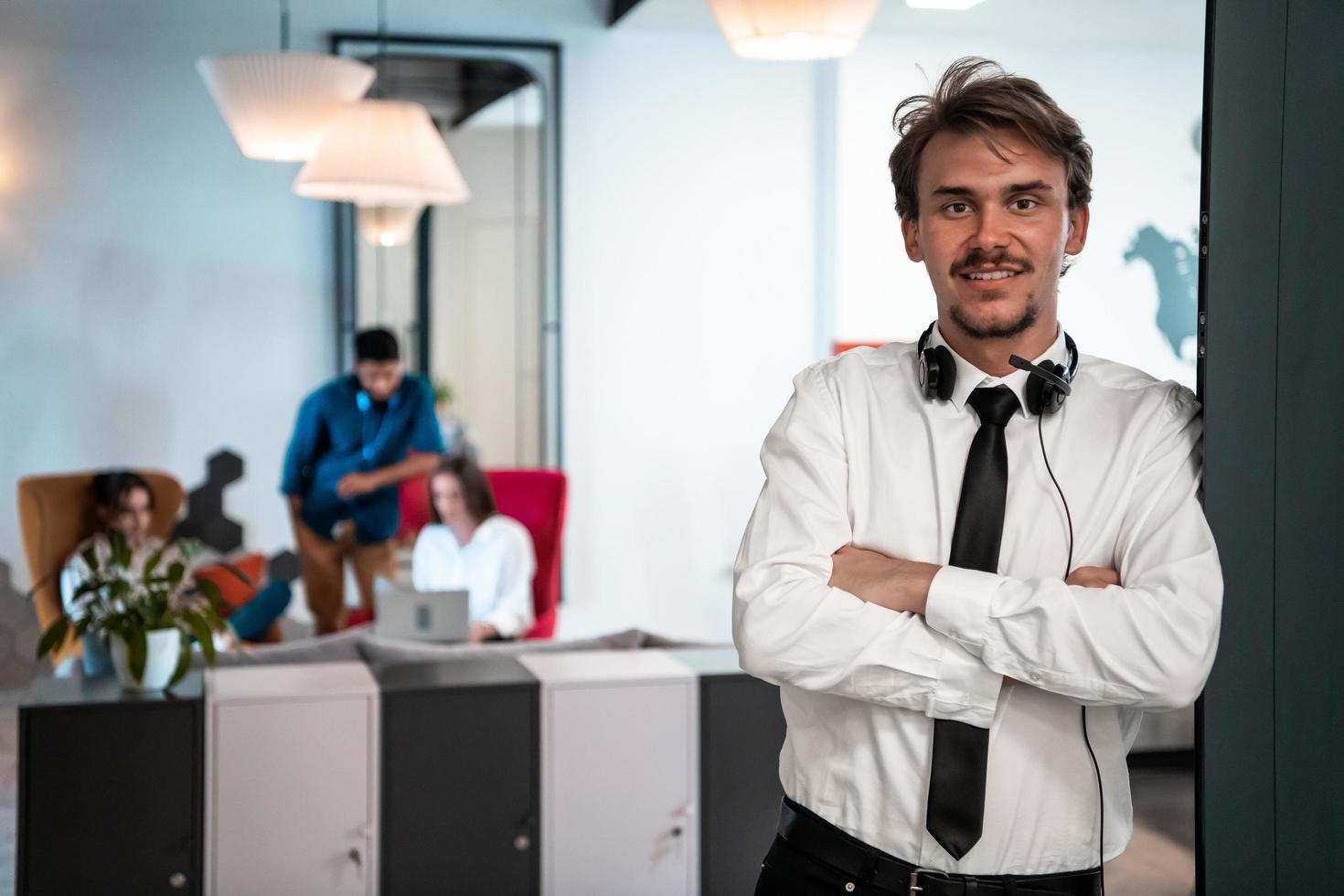 Portrait of successful businessman entrepreneur with headphones around his neck and colleagues in the background at busy startup office photo