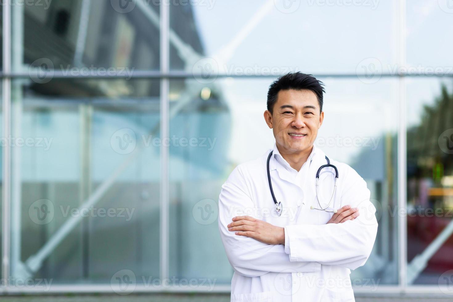 retrato de un alegre médico asiático sonriendo con las manos dobladas en el fondo de una clínica moderna foto