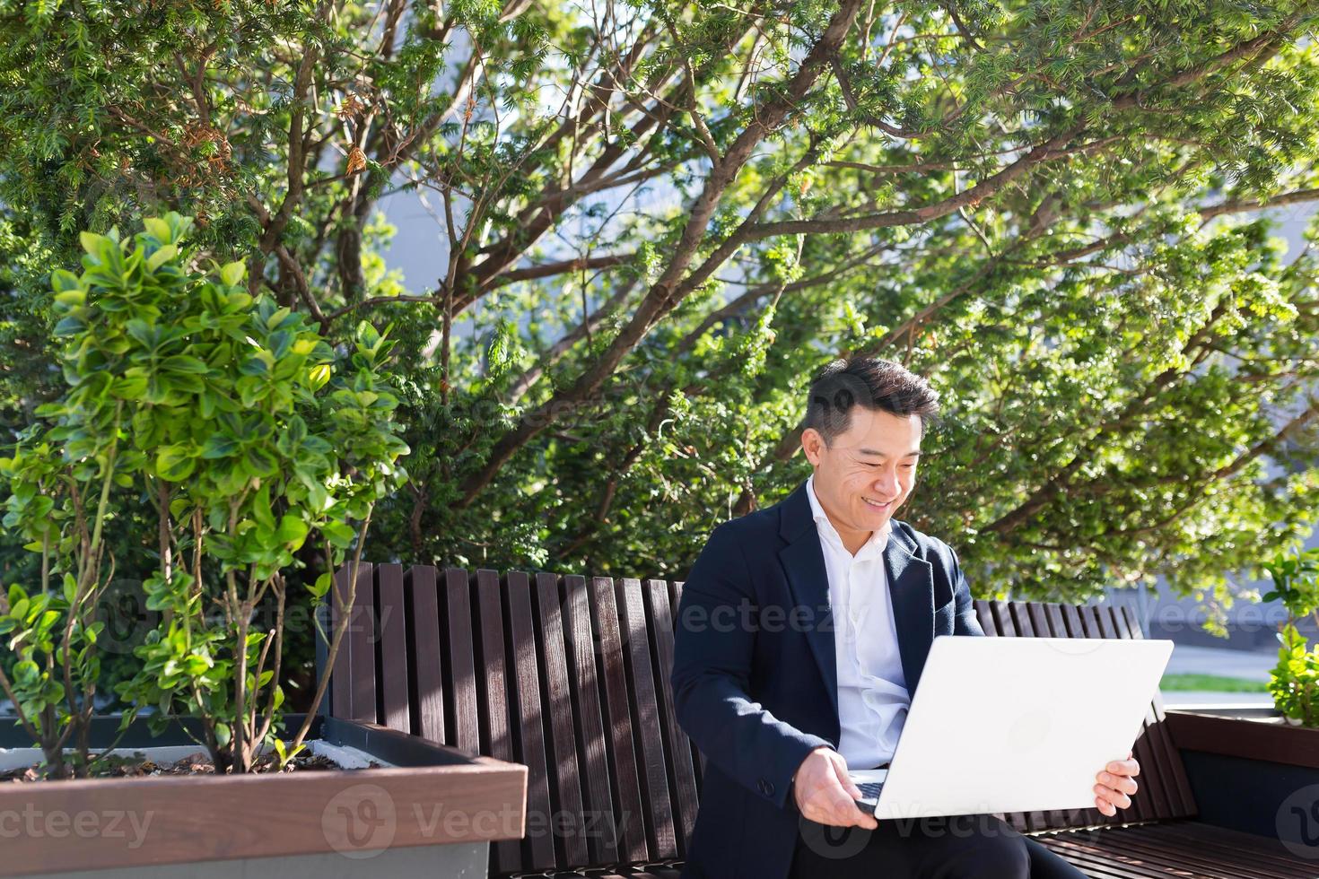 young asian business man or freelancer sitting on bench and working with laptop in city park photo