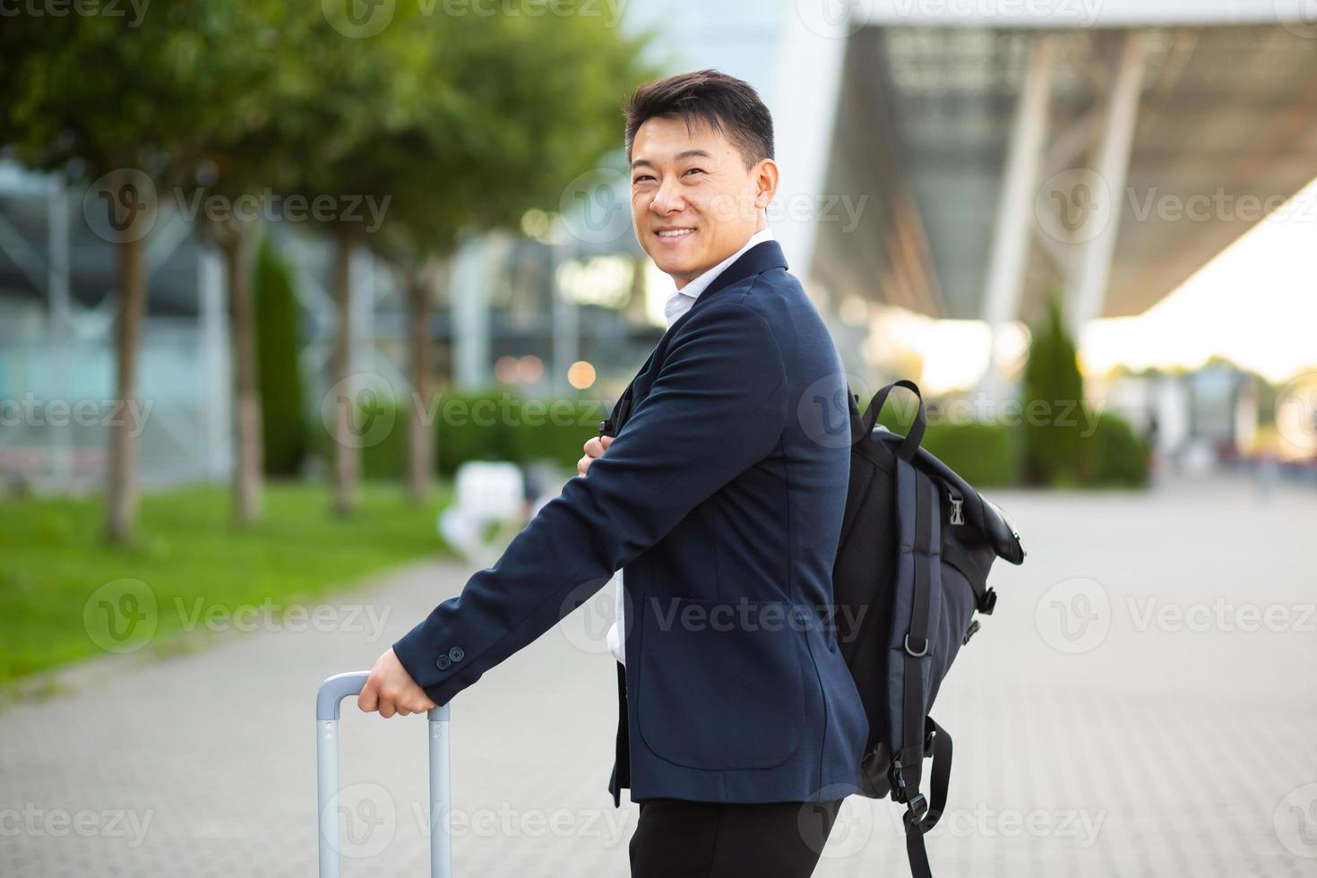 feliz turista asiático con una maleta grande cerca del aeropuerto, en una reunión de negocios llega buscando foto