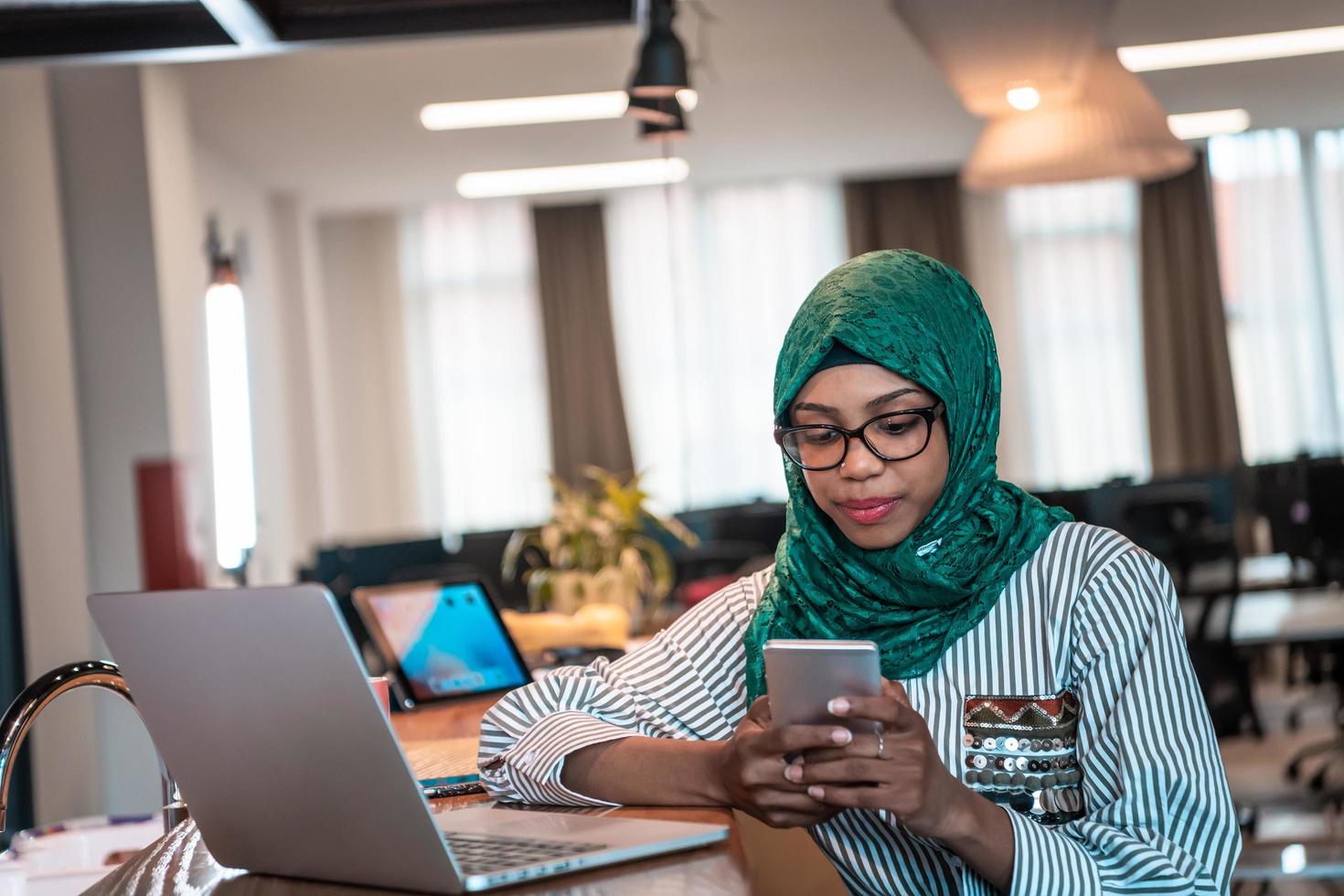 Smiling young woman talk on smartphone work on laptop look in distance thinking. Happy African American customer client use modern gadgets cellphone and computer at home office. photo