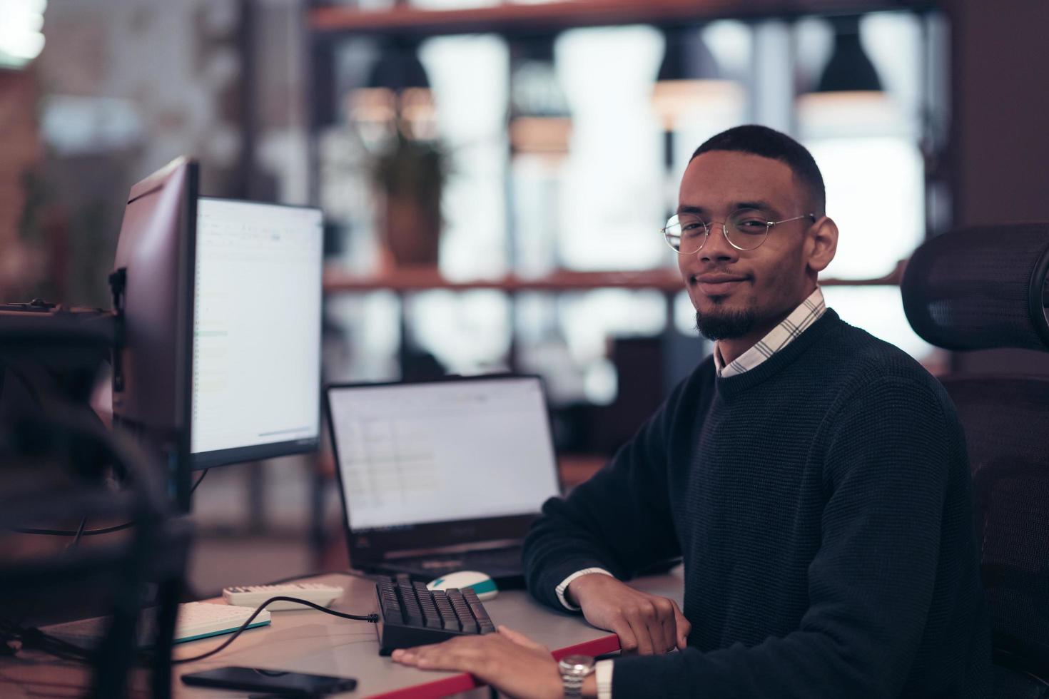 Young African American with glasses sitting in a modern coworking office and working at a computer photo