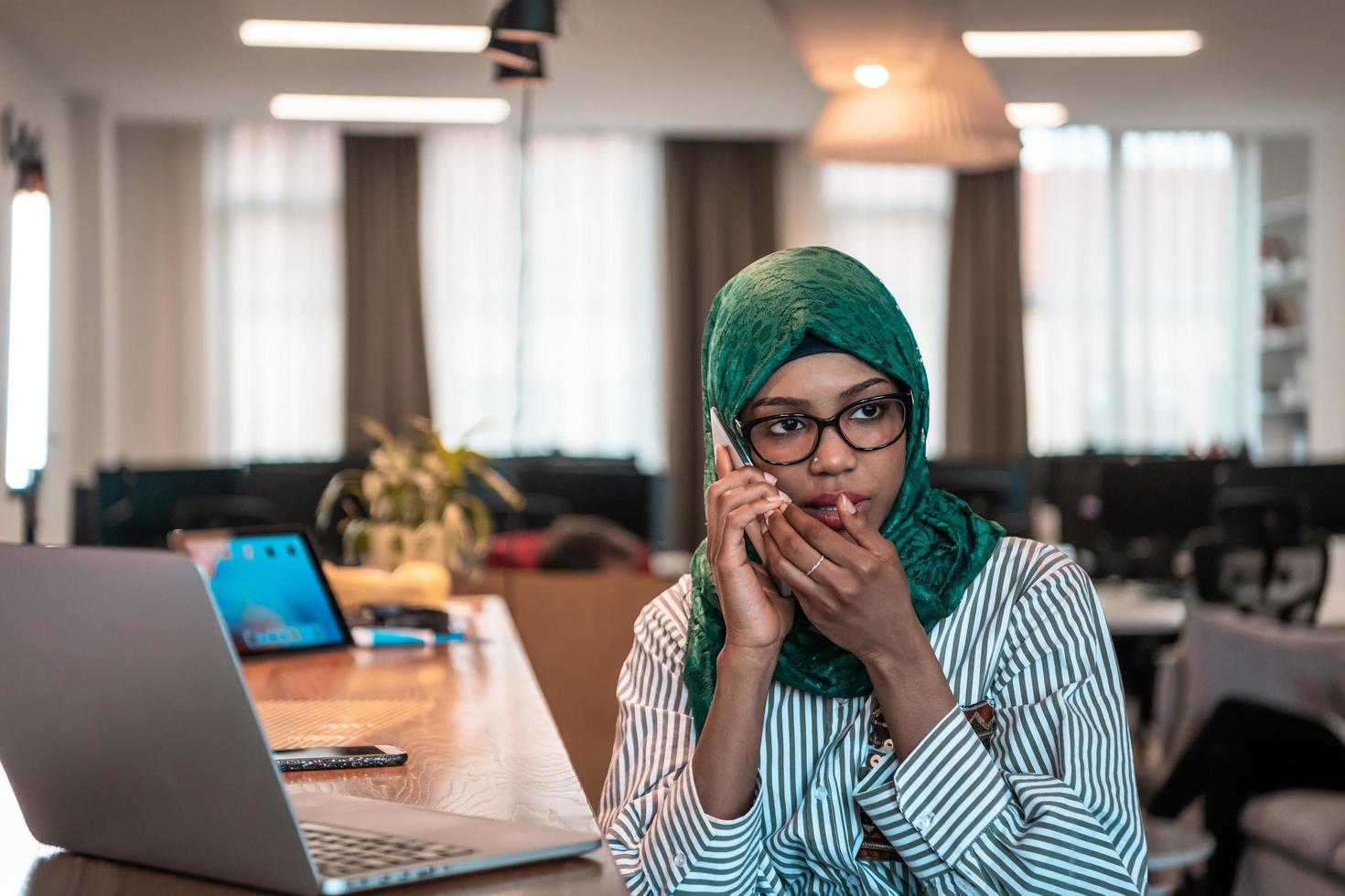 Smiling young woman talk on smartphone work on laptop look in distance thinking. Happy African American customer client use modern gadgets cellphone and computer at home office. photo