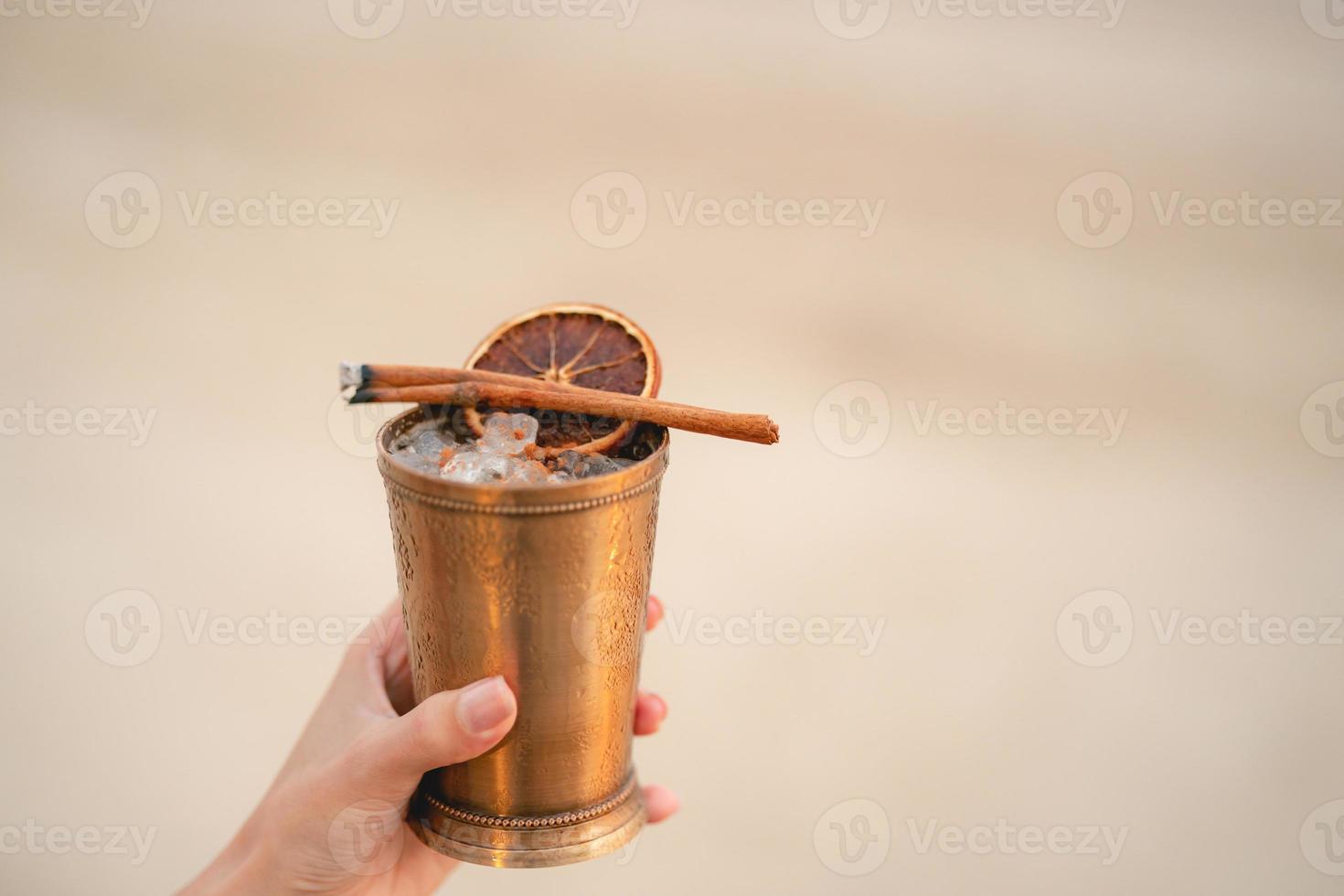 Hand with cocktail, Alcoholic drink with ice, orange and cinnamon on the beach. photo