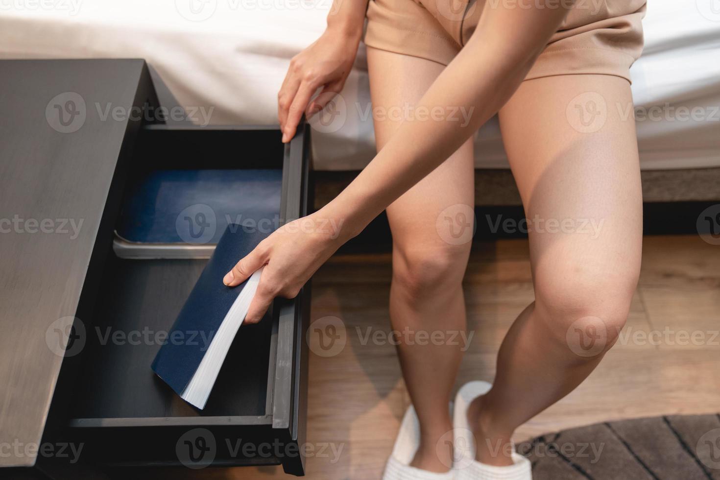 Woman sitting on bed with white sheet and next to wooden drawer stand and note book inside. photo