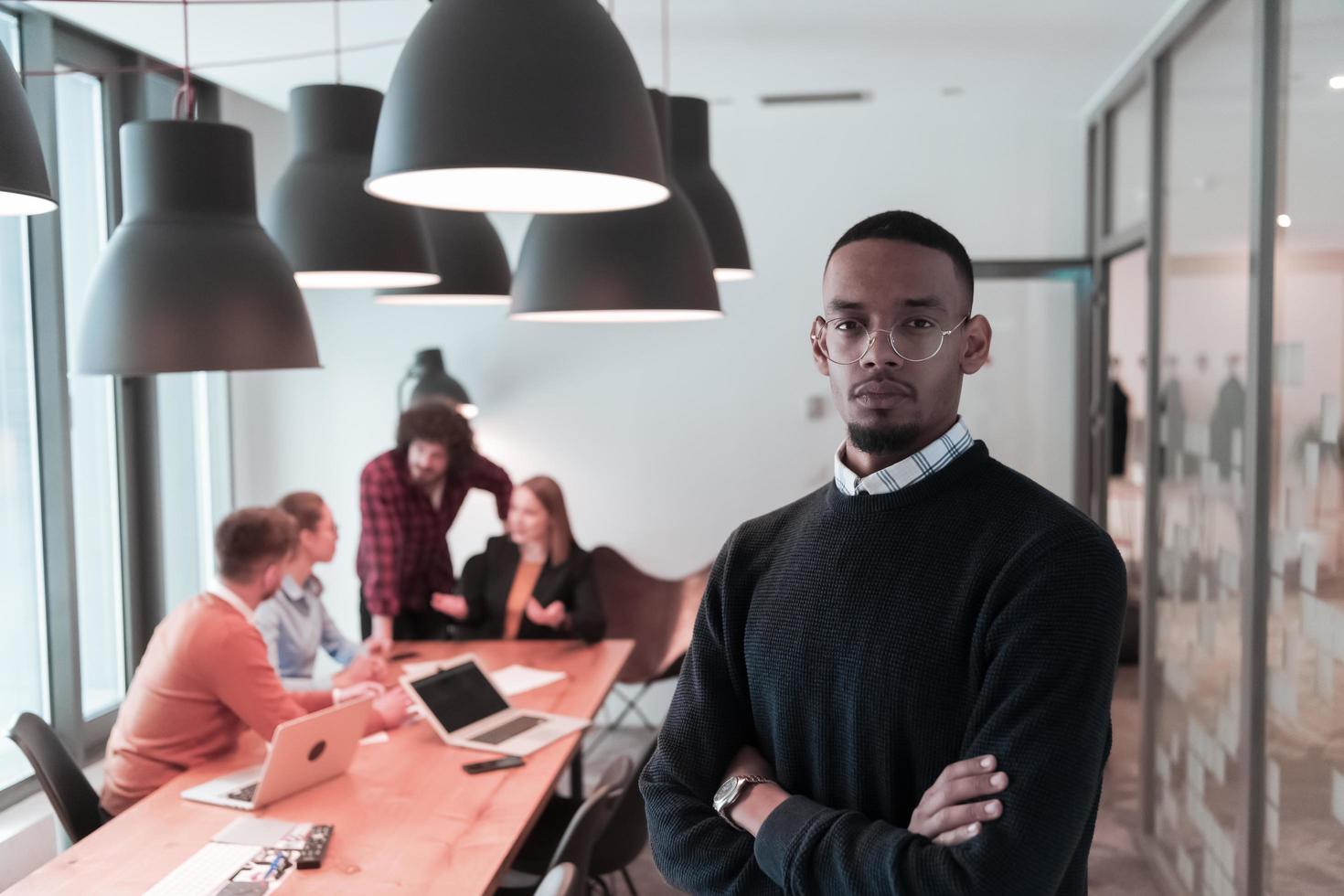 Portrait of happy millennial male business owner in modern office. Businessman wearing glasses, smiling and looking at camera. Busy diverse team working in background. Leadership concept. Head shot. photo