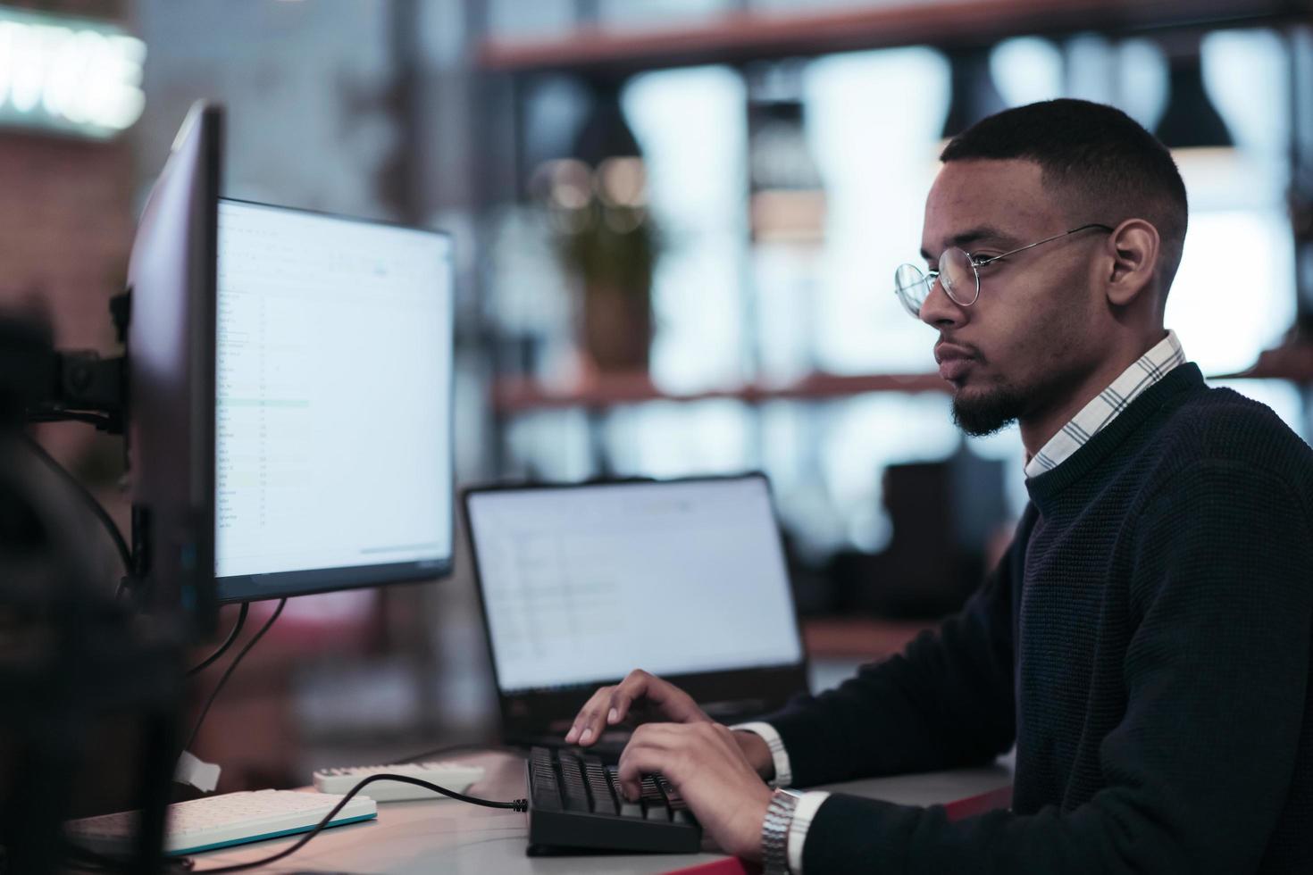 Young African American with glasses sitting in a modern coworking office and working at a computer photo