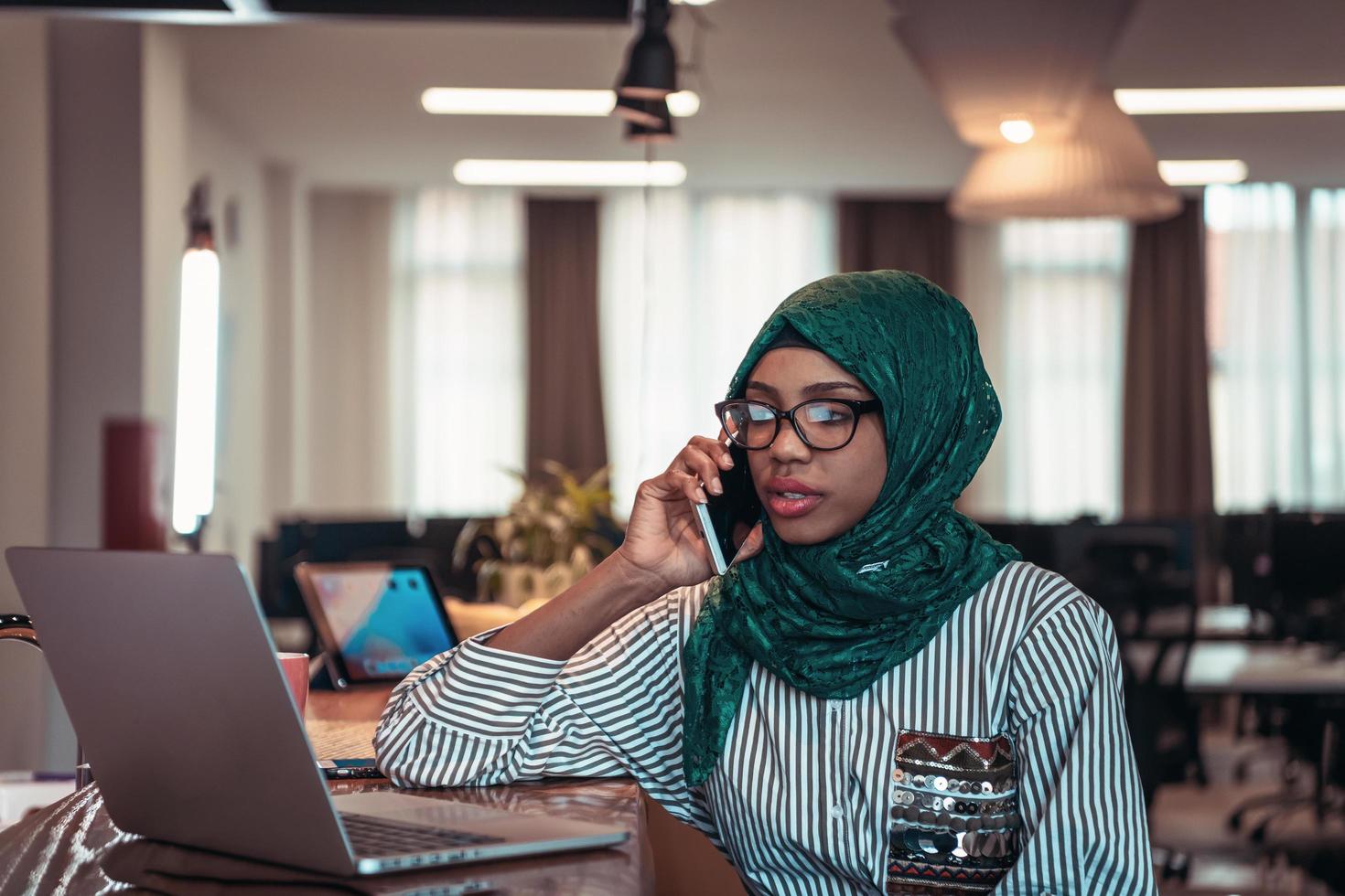 Smiling young woman talk on smartphone work on laptop look in distance thinking. Happy African American customer client use modern gadgets cellphone and computer at home office. photo