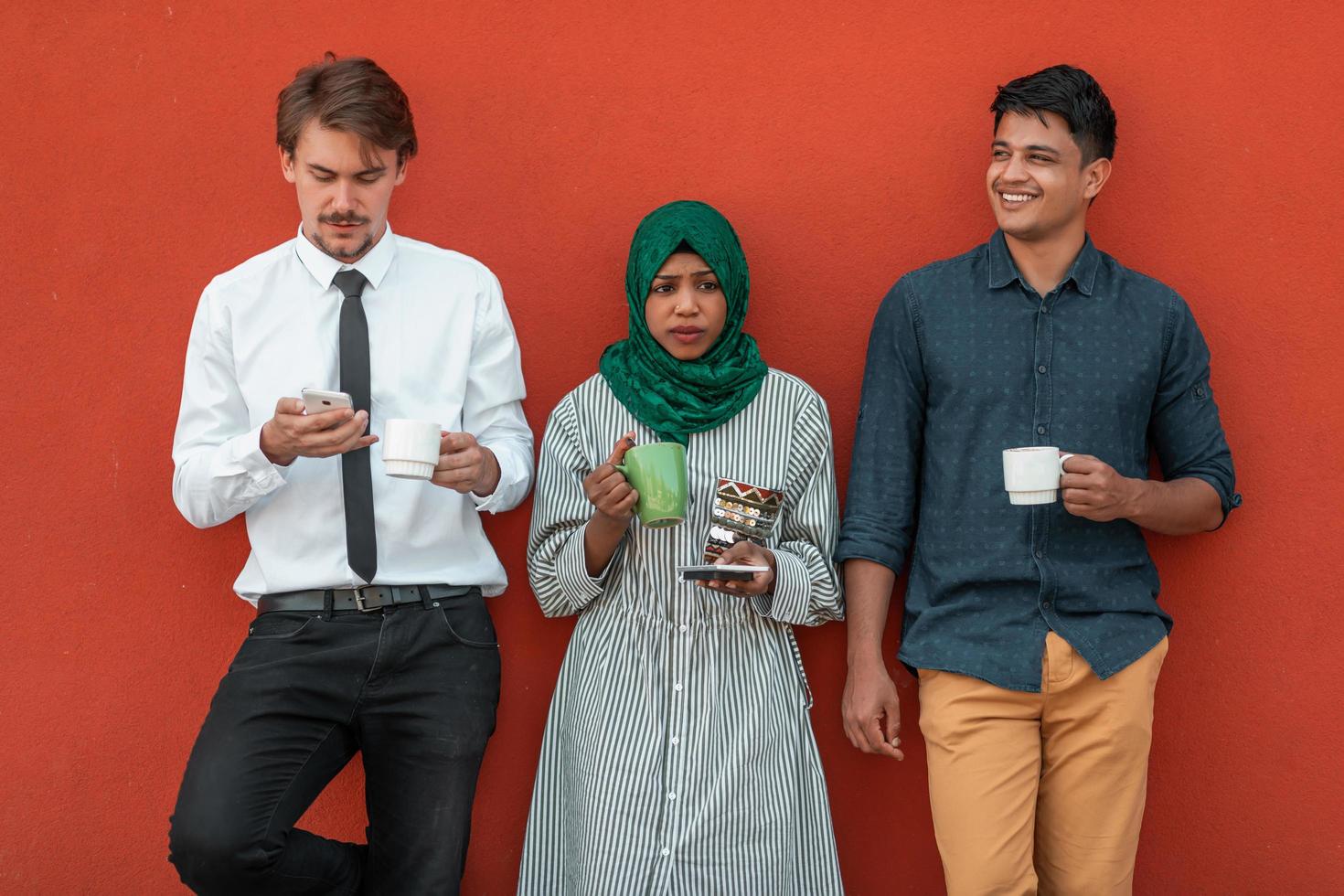Multiethnic group of casual businesspeople using smartphone during a coffee break from work in front of the red wall outside. photo