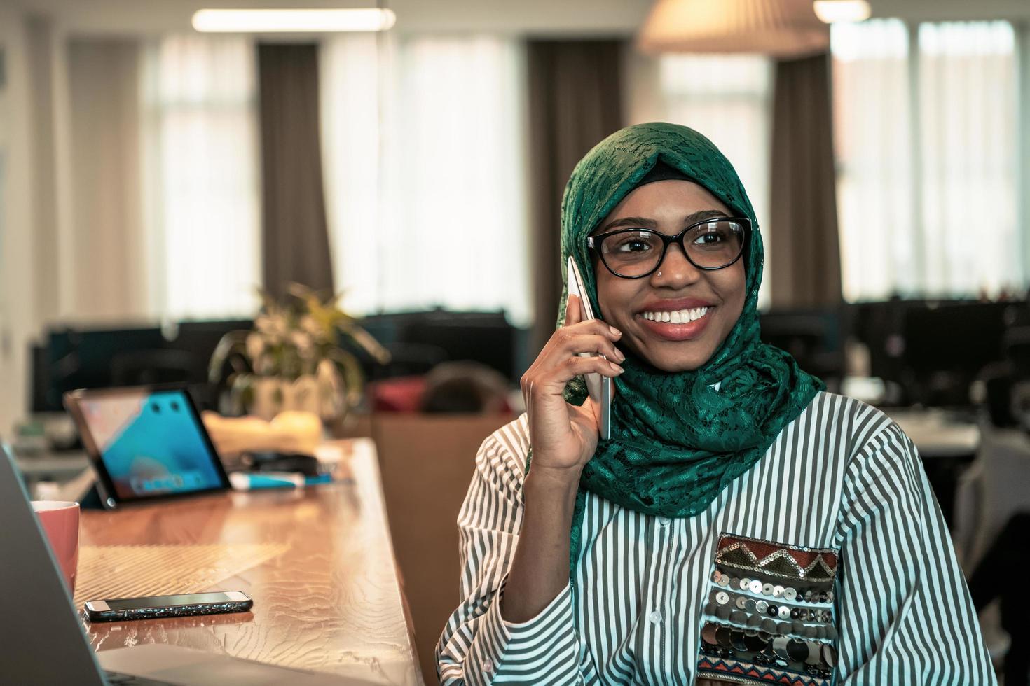 Smiling young woman talk on smartphone work on laptop look in distance thinking. Happy African American customer client use modern gadgets cellphone and computer at home office. photo