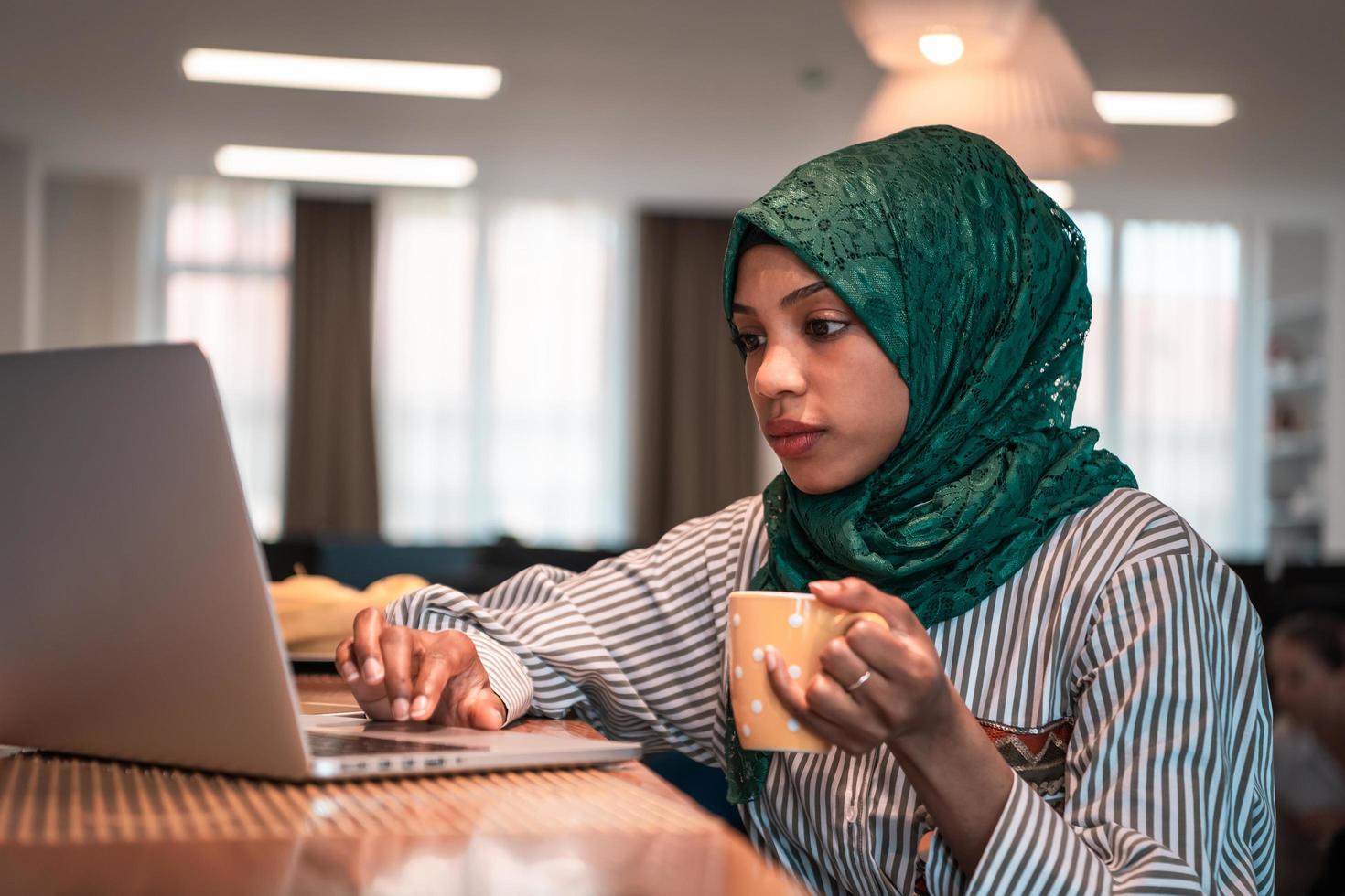 mujer de negocios musulmana africana que usa un hiyab verde bebiendo té mientras trabaja en una computadora portátil en el área de relajación en la moderna oficina de inicio de planta abierta. foto