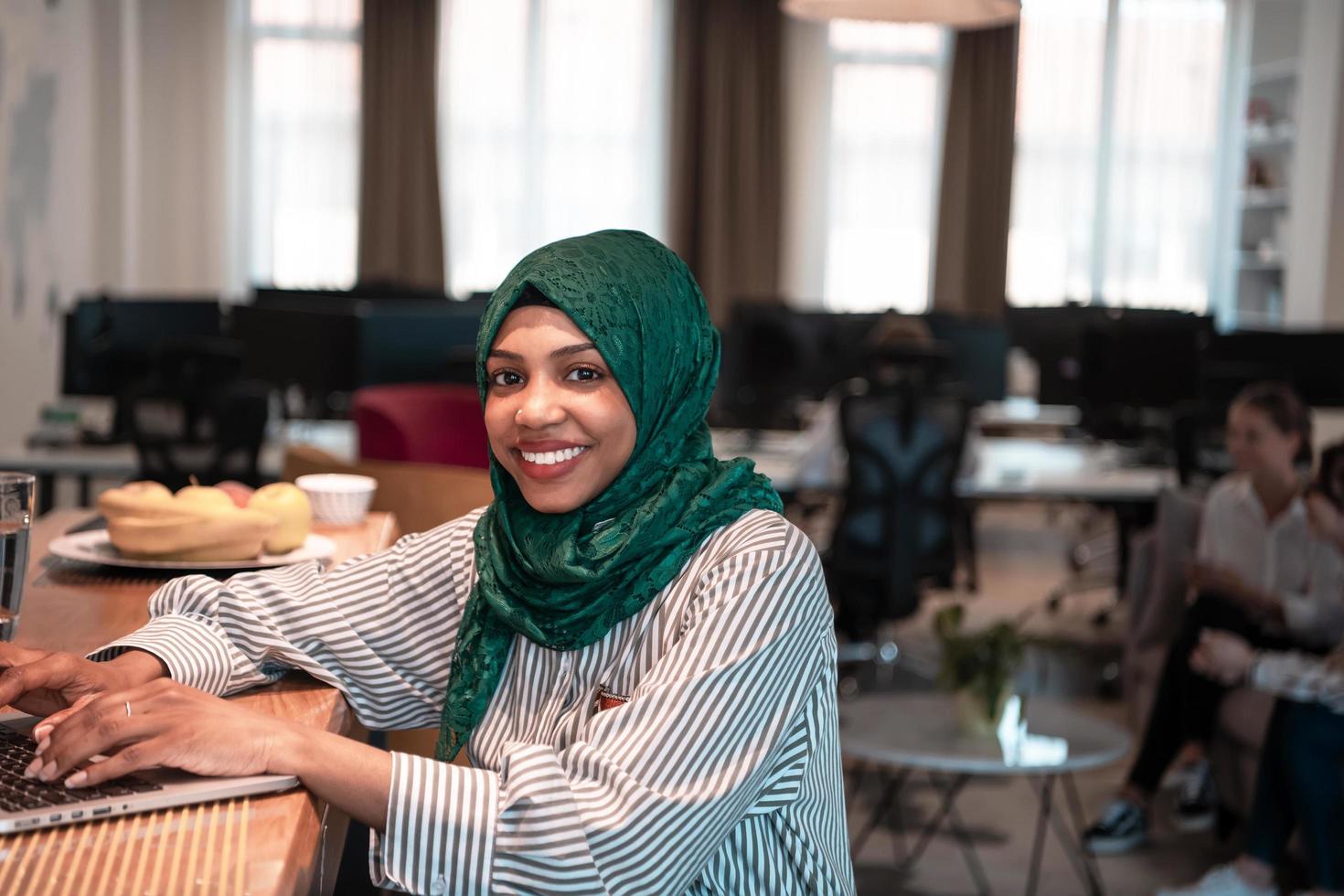 mujer de negocios musulmana africana que usa un hiyab verde y trabaja en una computadora portátil en el área de relajación en la oficina de inicio moderna de planta abierta. foto