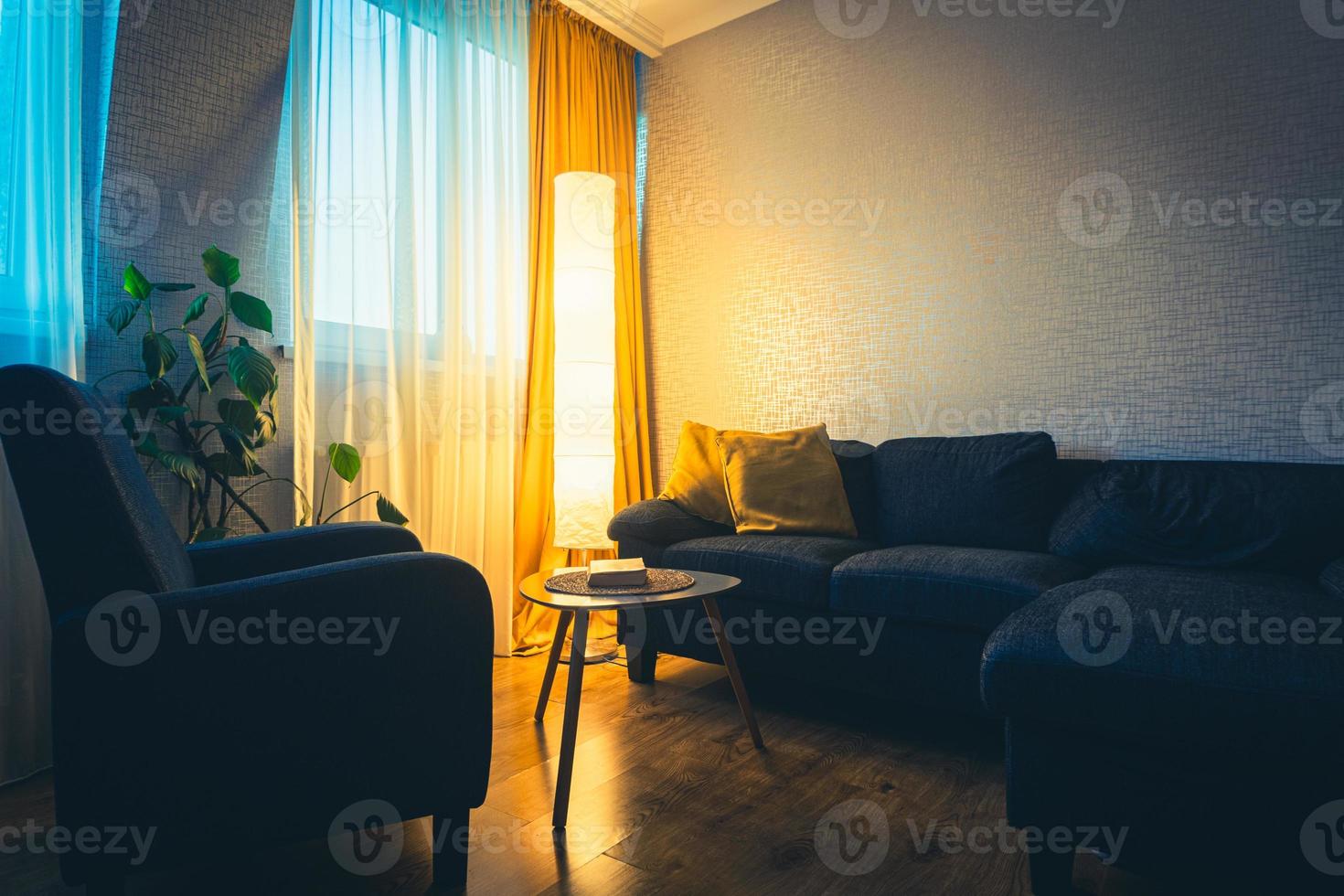 Cozy apartment living room with sofa and yellow pillows and stylisg table with book by flower and window with blue light outside in winter. Early evening book read. Copy paste wall photo