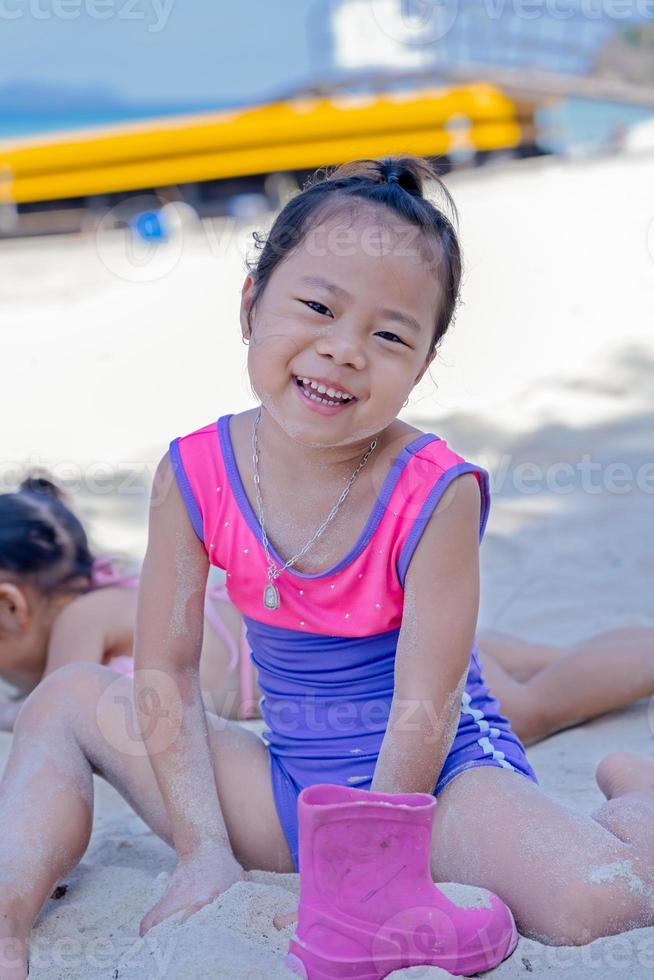 Close-up of adorable happy child girl playing with sand on ocean beach during summer tropical vacations. Cute Asian little child with a swimsuit laughing and having fun playing with sand on the beach photo
