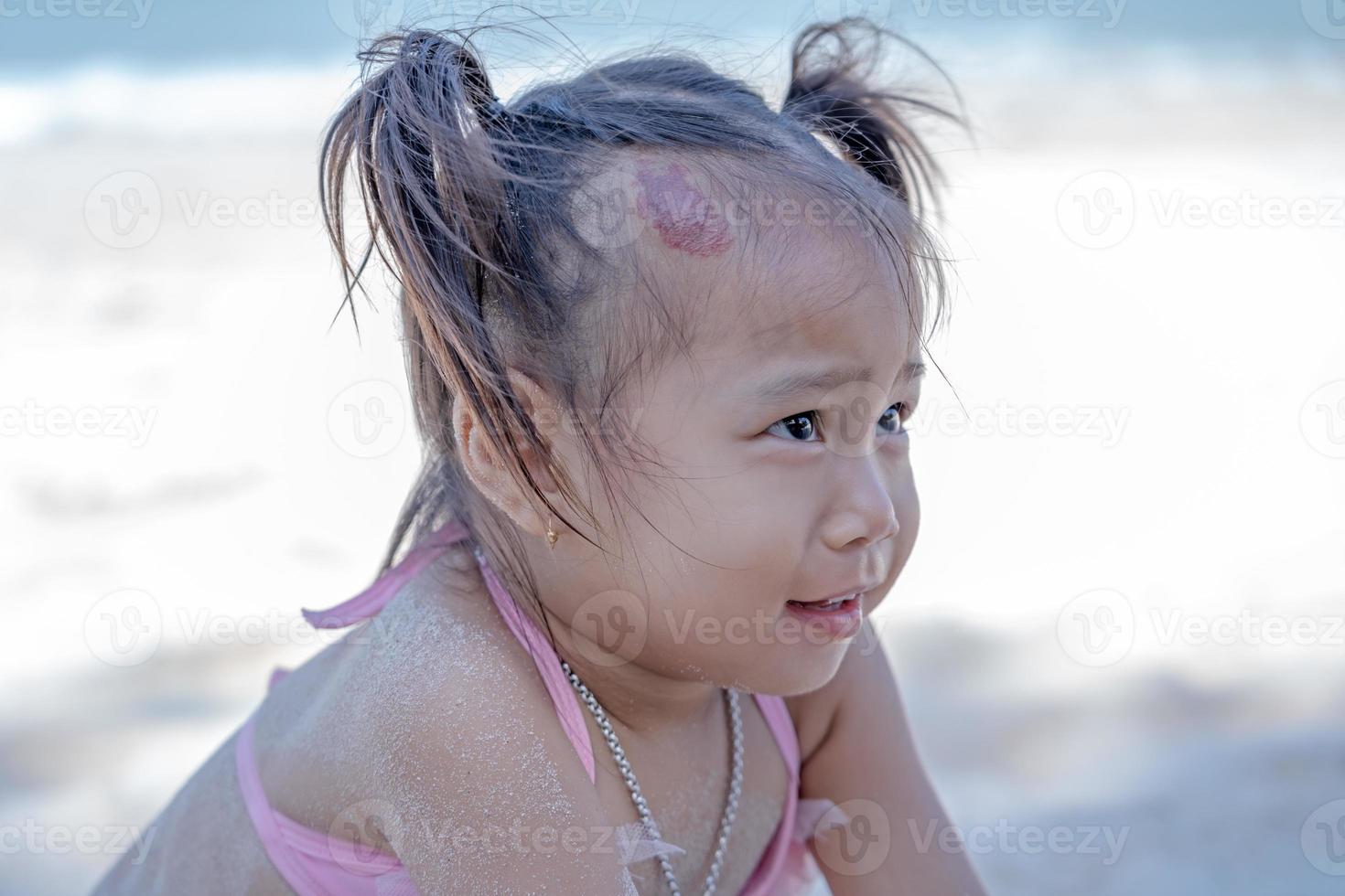 Adorable happy child girl playing with sand on ocean beach during summer tropical vacations. Funny face little child looking mother with big capillary strawberry hemangiomas red birthmark on head photo