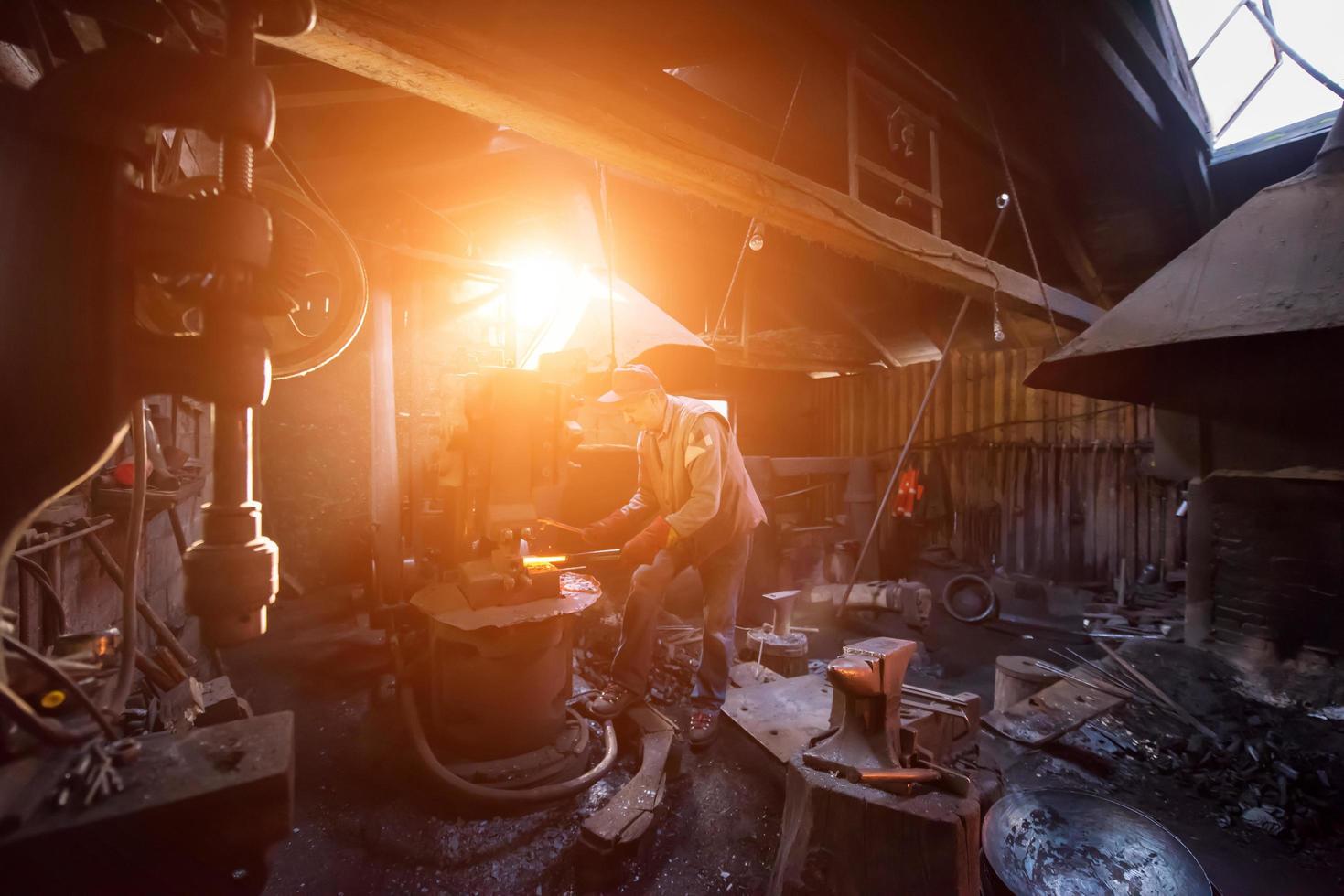 herrero forja manualmente el metal fundido con la luz del sol a través de las ventanas foto