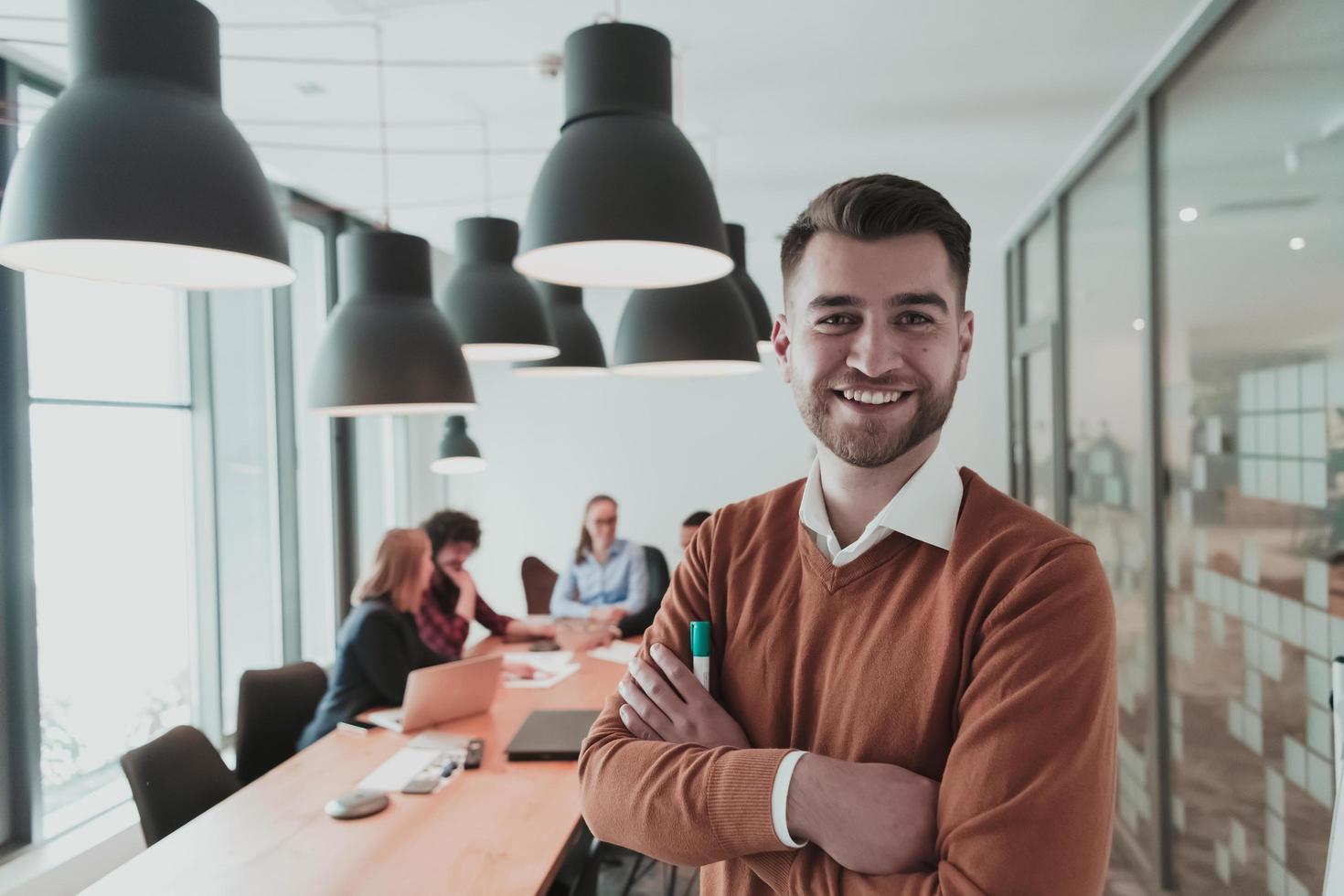 Portrait of happy millennial male business owner in modern office. Businessman wearing glasses, smiling and looking at camera. Busy diverse team working in background. Leadership concept. Head shot. photo