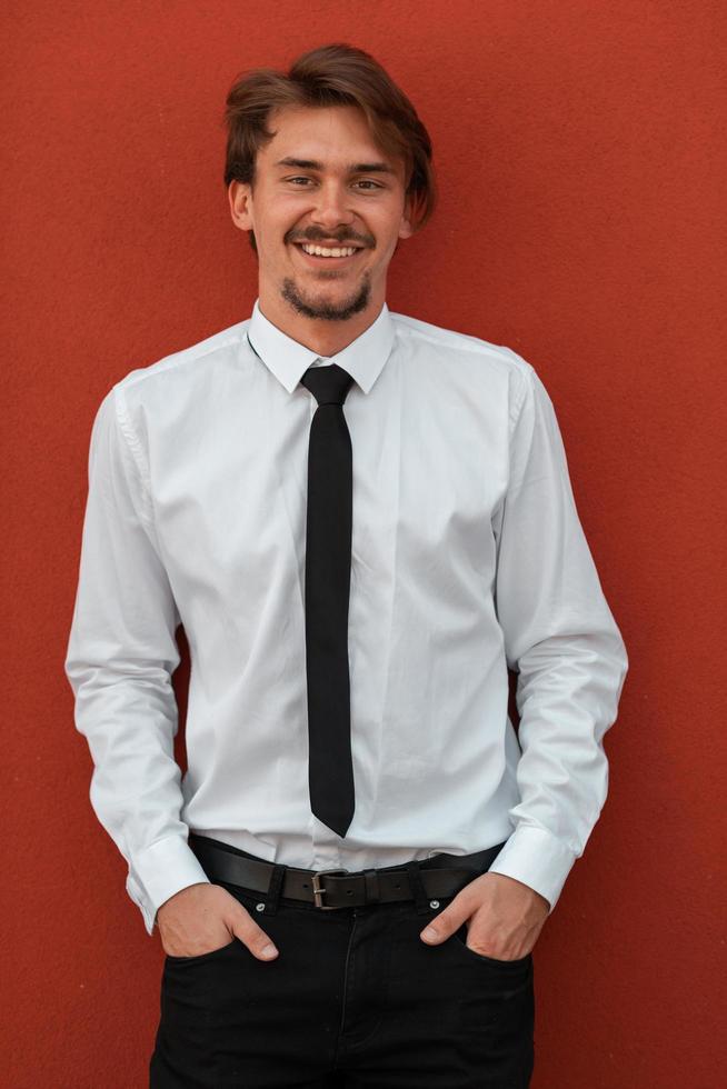 retrato de un nuevo hombre de negocios con una camisa blanca y una corbata negra parado frente a una pared roja afuera foto