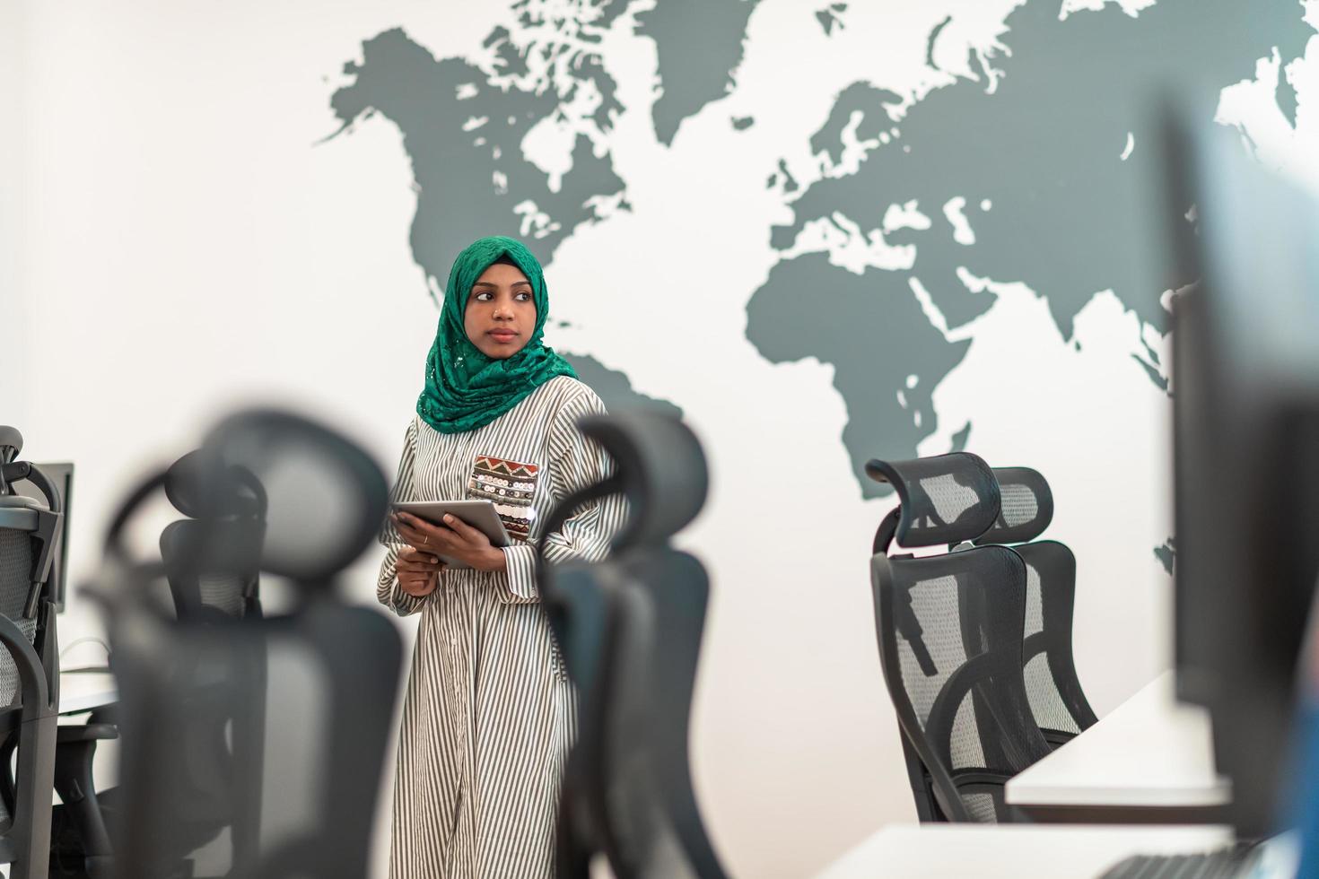 Portrait of muslim female software developer with green hijab holding tablet computer while standing at modern open plan startup office photo