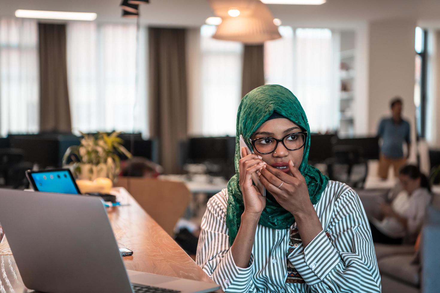 Smiling young woman talk on smartphone work on laptop look in distance thinking. Happy African American customer client use modern gadgets cellphone and computer at home office. photo