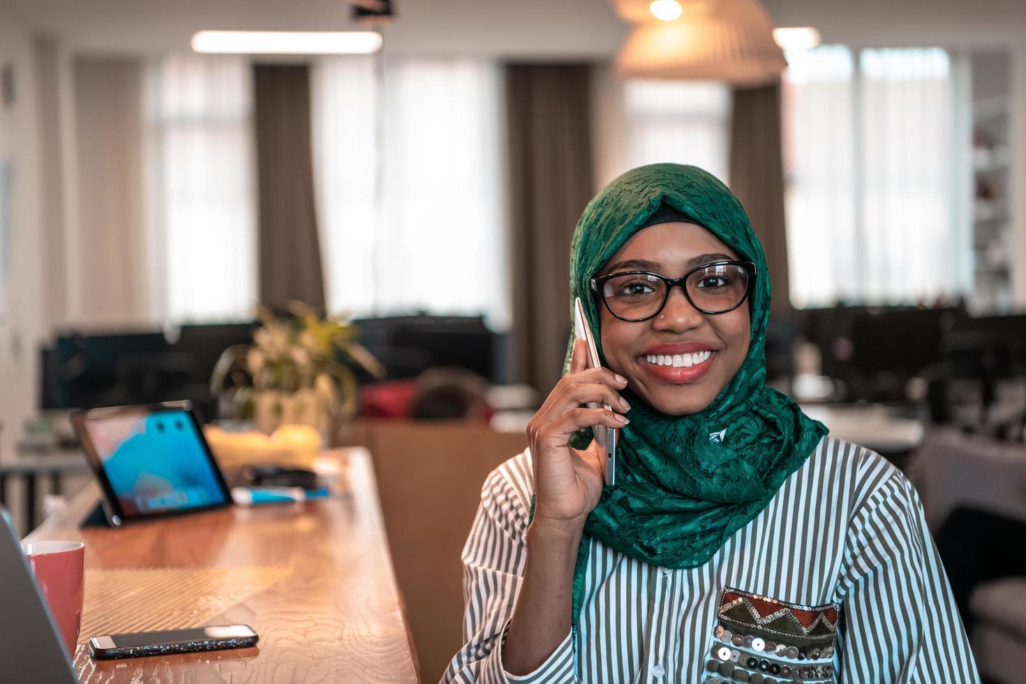 Smiling young woman talk on smartphone work on laptop look in distance thinking. Happy African American customer client use modern gadgets cellphone and computer at home office. photo