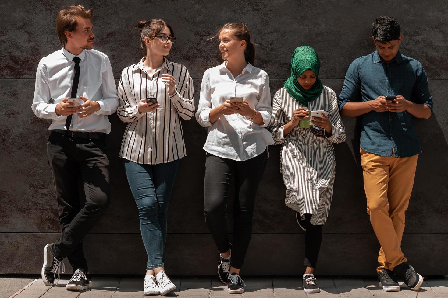 Multiethnic group of casual business people using mobile phone during coffee break from work in front of black wall outside photo