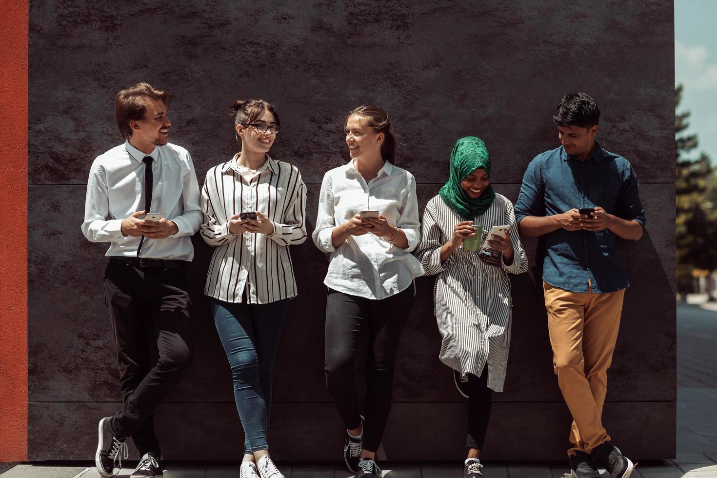 Multiethnic group of casual business people using mobile phone during coffee break from work in front of black wall outside photo