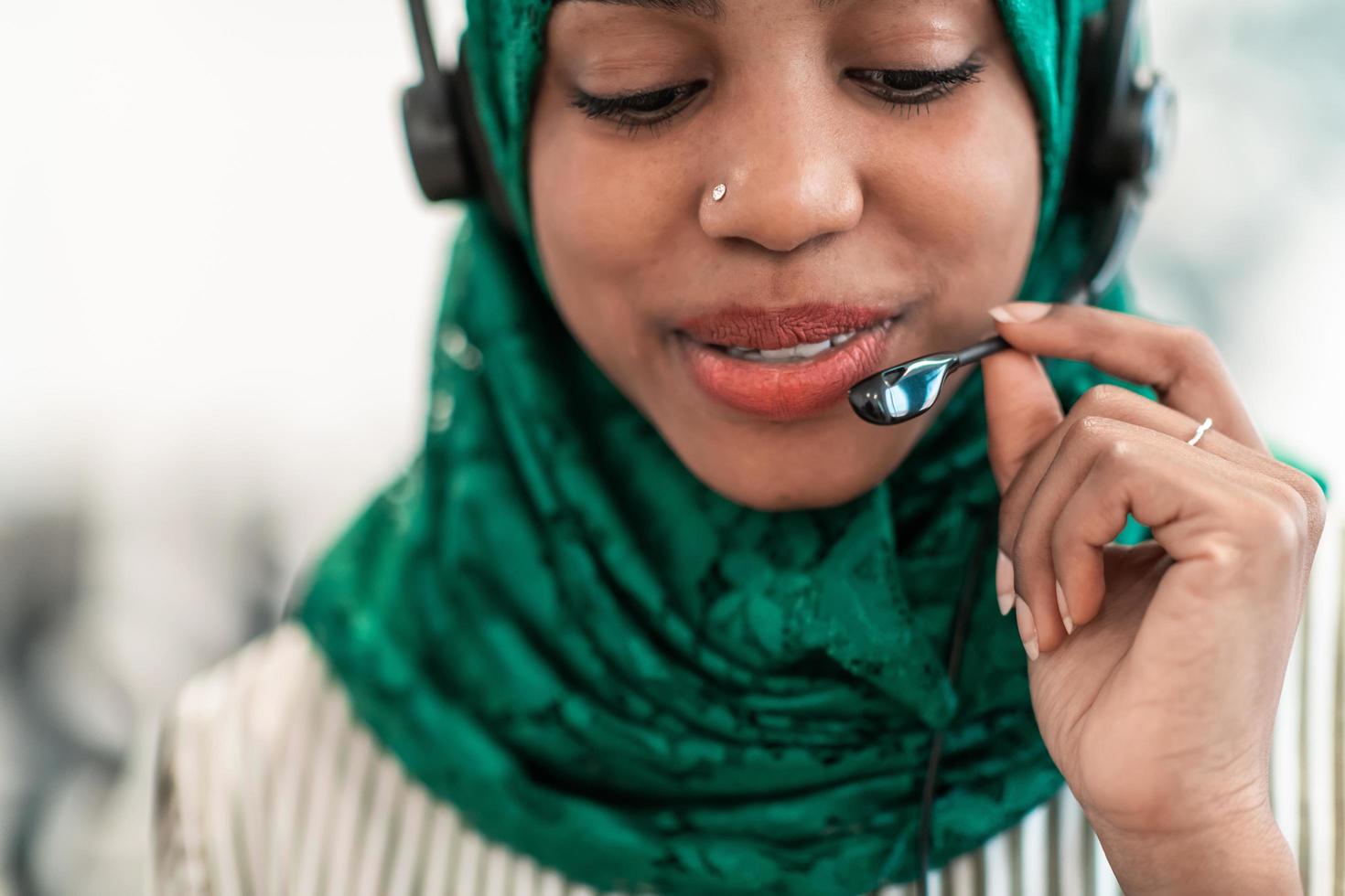 Afro Muslim female with green hijab scarf customer representative business woman with phone headset helping and supporting online with customer in modern call centre photo