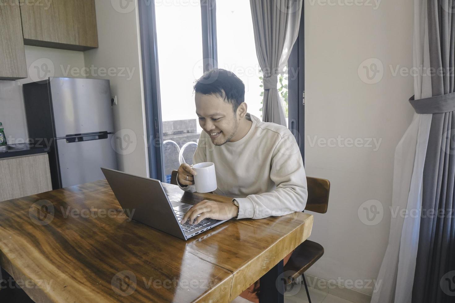 un joven asiático pasa su tiempo en casa sentado en el comedor trabajando felizmente en su computadora portátil mientras sostiene una taza foto