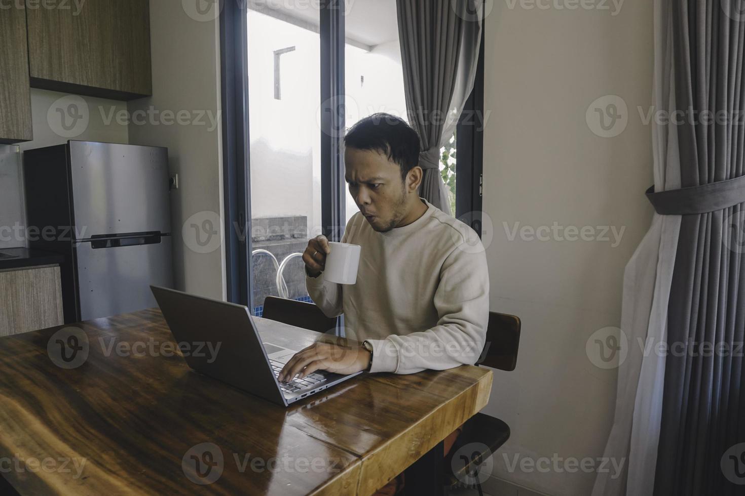un joven asiático pasa su tiempo en casa sentado en el comedor conmocionado mientras trabaja en su computadora portátil mientras sostiene una taza foto