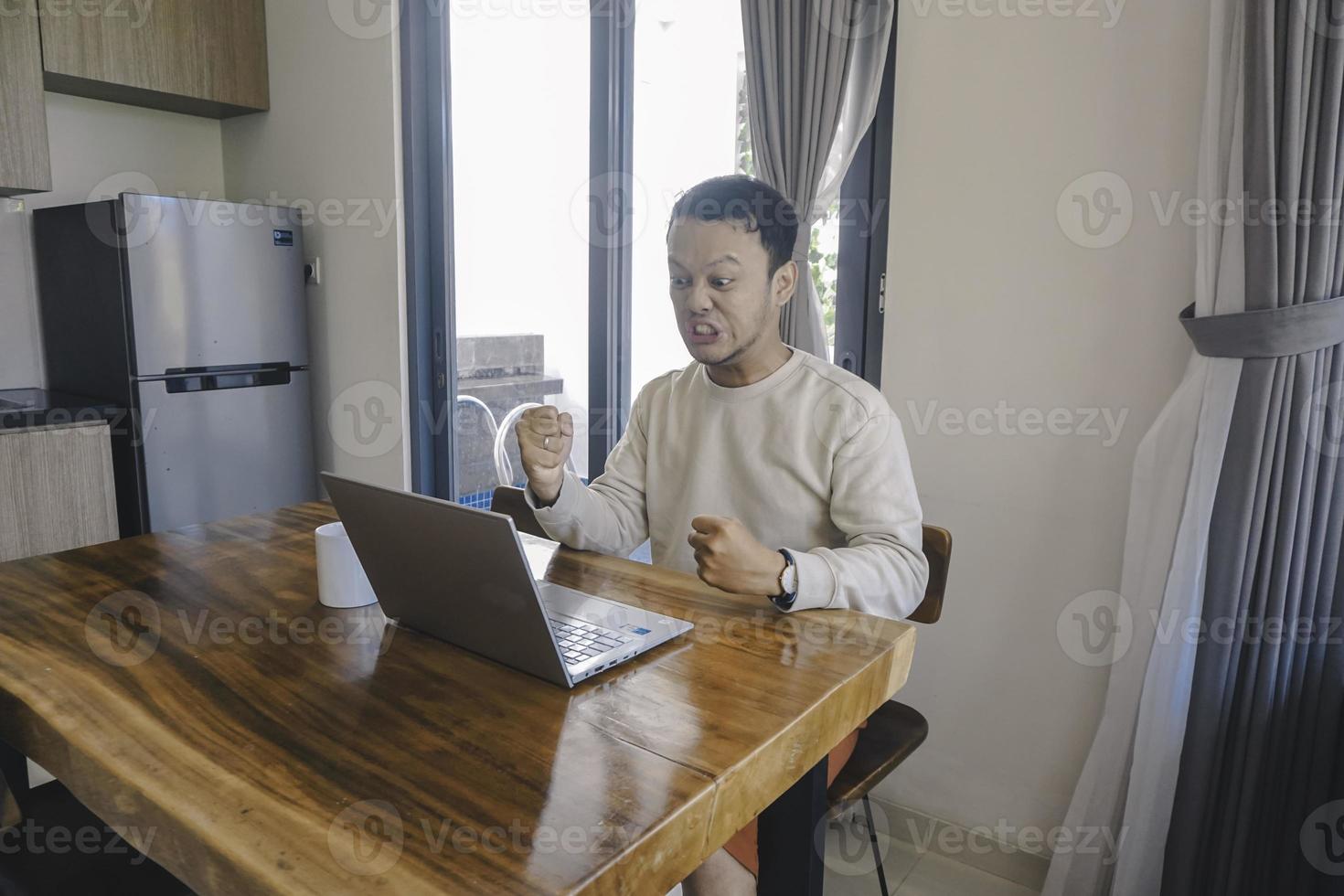 un joven asiático pasa su tiempo en casa sentado en el comedor trabajando en su computadora portátil y se siente frustrado foto