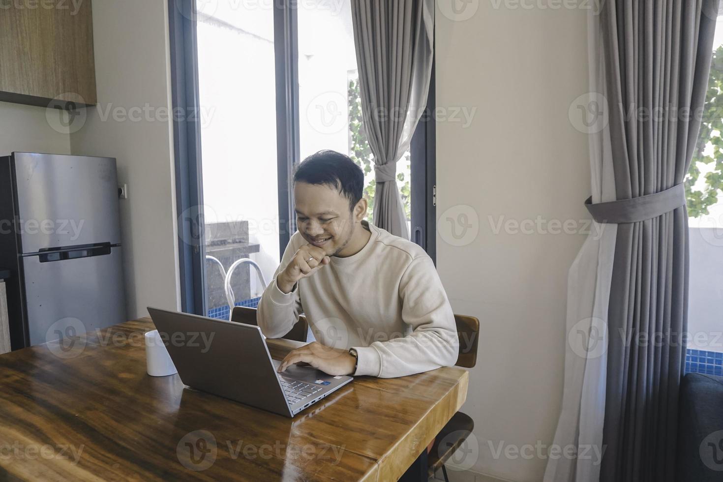 un joven asiático pasa su tiempo en casa sentado en el comedor trabajando felizmente en su computadora portátil foto