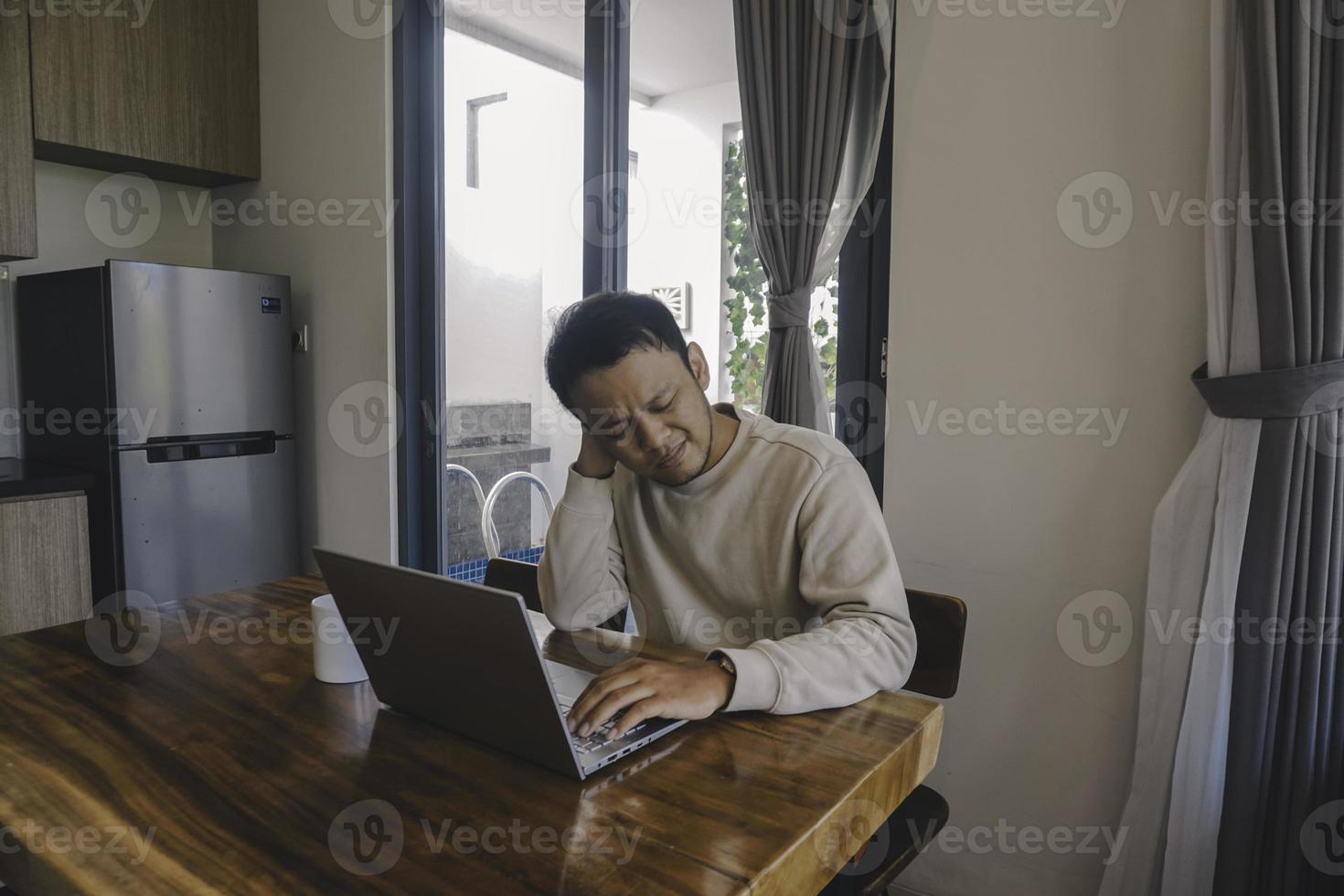 un joven asiático pasa su tiempo en casa sentado en el comedor trabajando en su computadora portátil y se siente frustrado foto