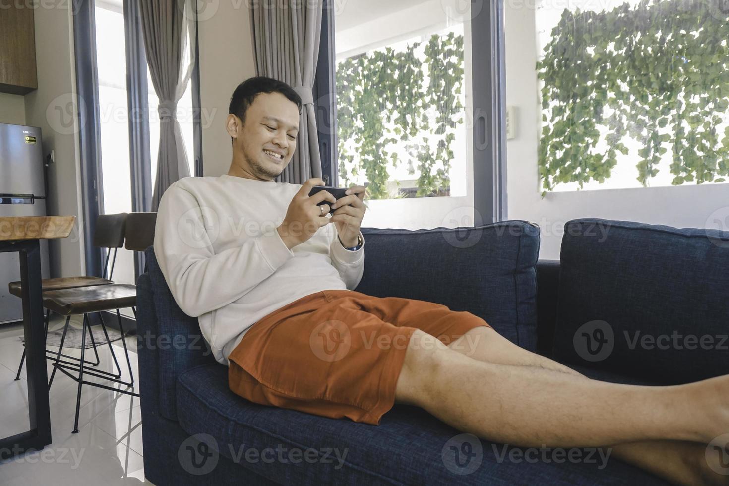 Young Asian man spend his free time at home sitting on a cozy sofa in the living room texting message on mobile app on his smartphone. photo
