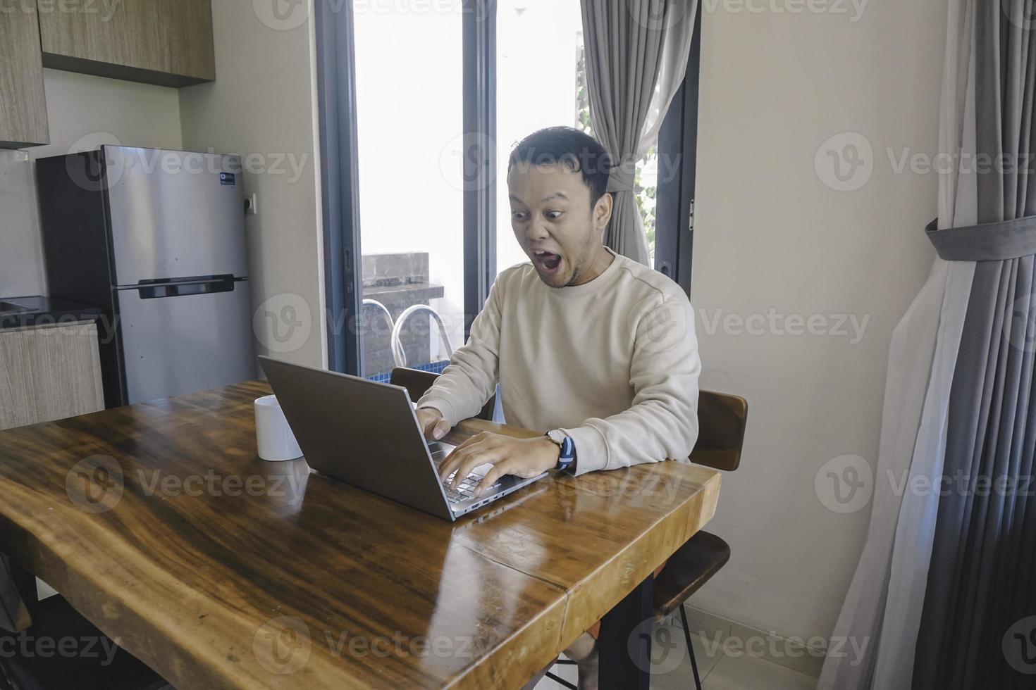 un joven asiático pasa su tiempo en casa sentado en el comedor conmocionado mientras mira su computadora portátil foto