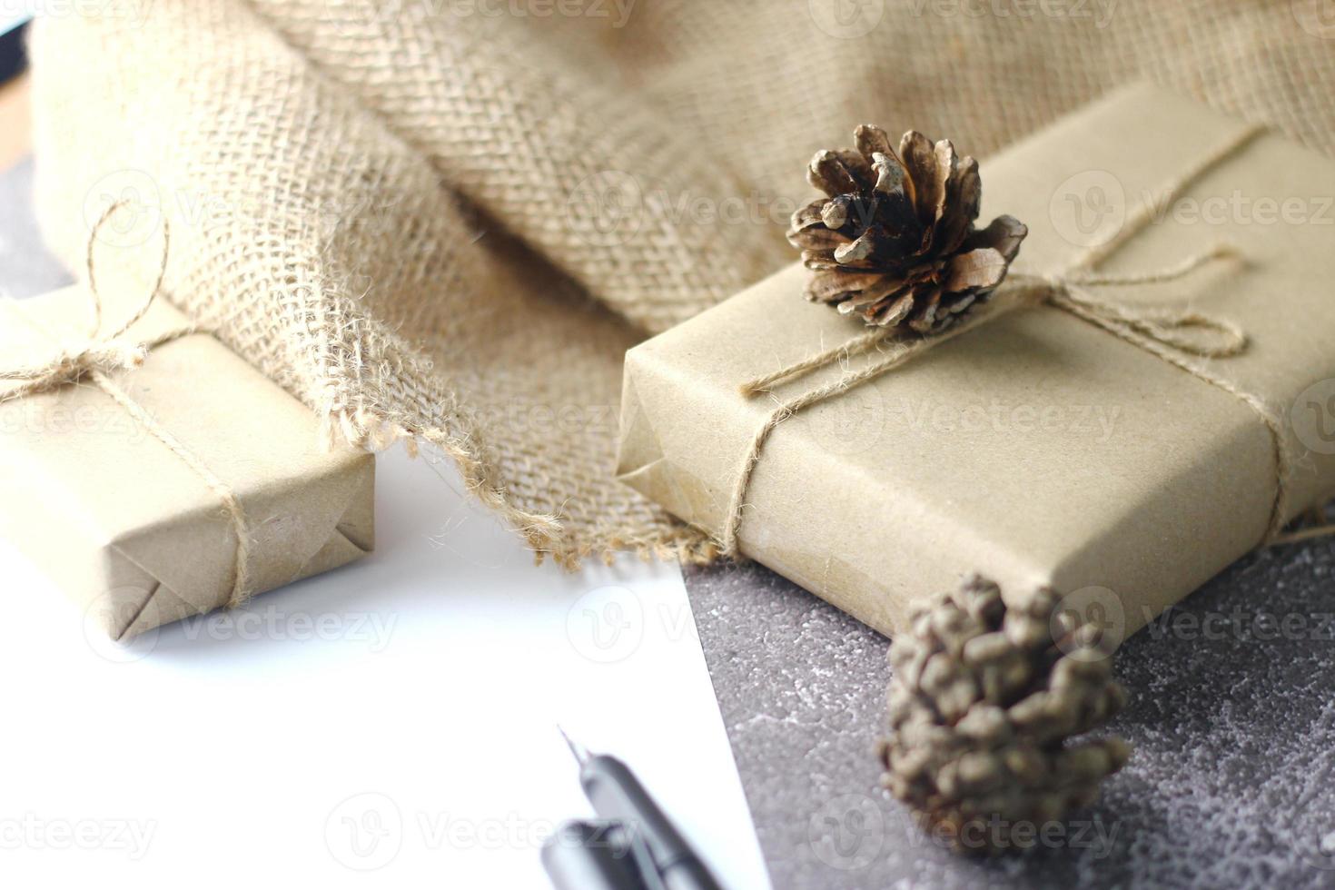 regalos de vacaciones caja de regalo de navidad y año nuevo envuelto en papel marrón, piñas y papel blanco y bolígrafos colocados en el espacio de copia de la mesa. foto