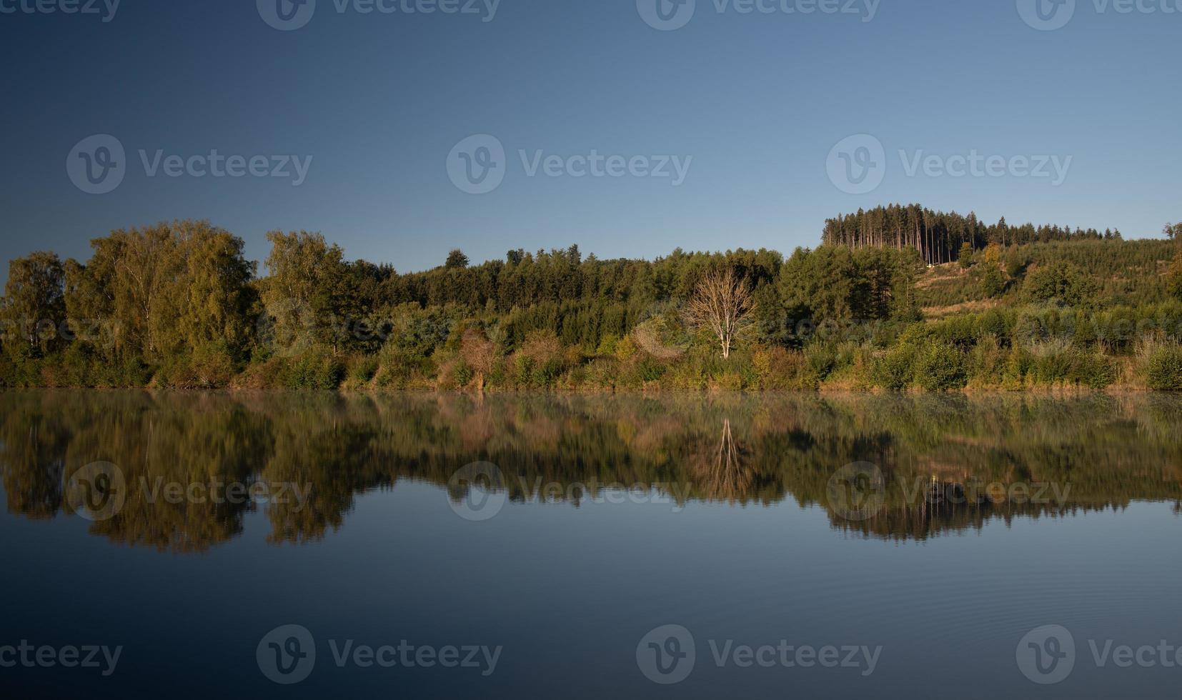 A forest is reflected in a lake in autumn, against a blue sky in Bavaria. photo