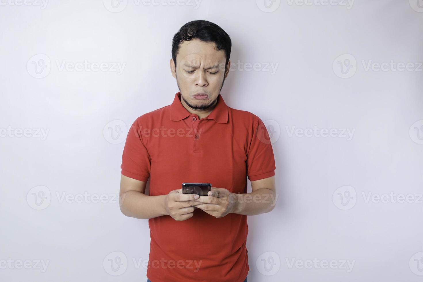 A dissatisfied young Asian man looks disgruntled wearing red t-shirt irritated face expressions holding his phone photo