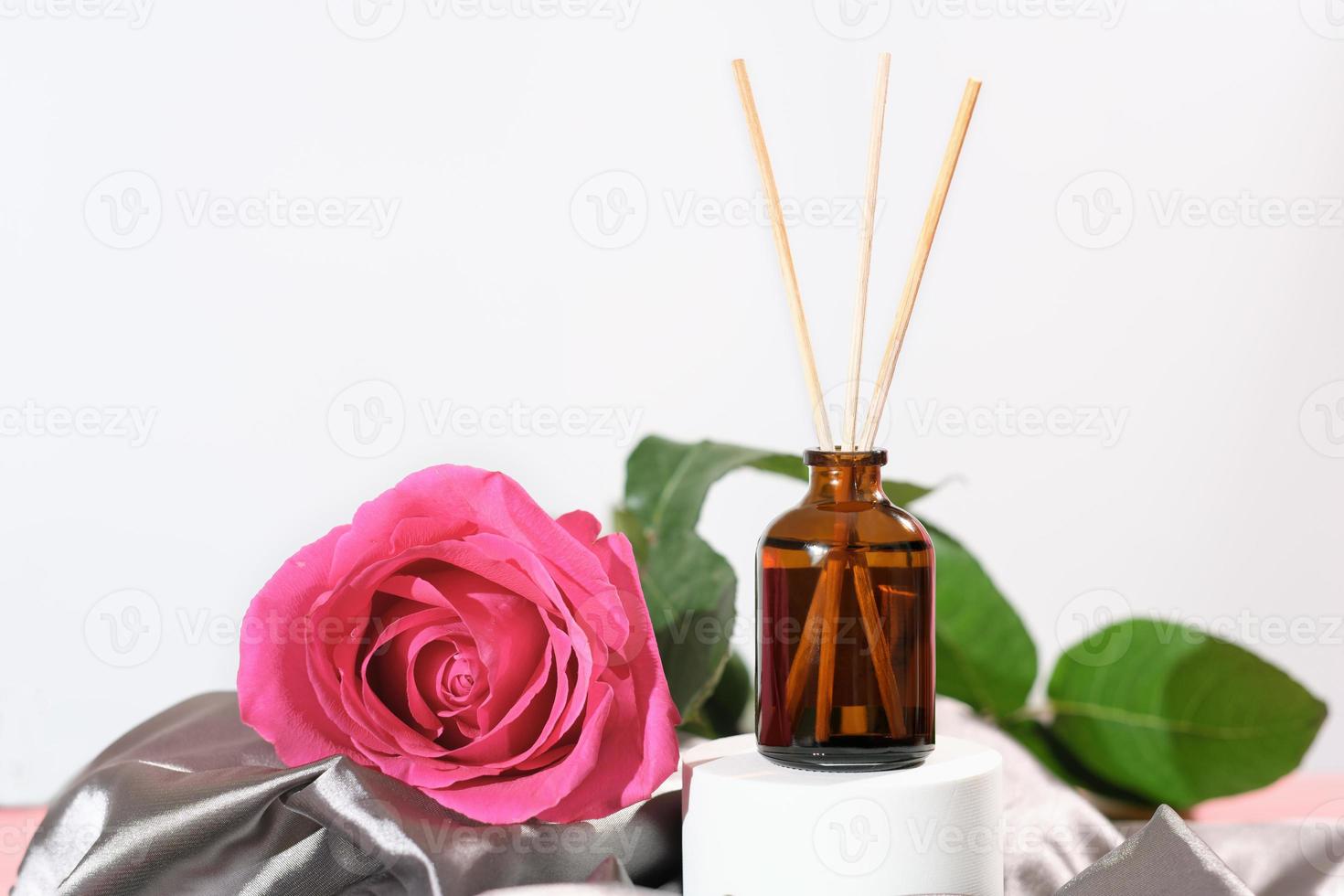 home aroma diffuser with reed sticks, rose flower scent. cozy home, romantic ambience, silk satin bedding sheets. pink rose flower next to glass bottle with fragranced oil. photo