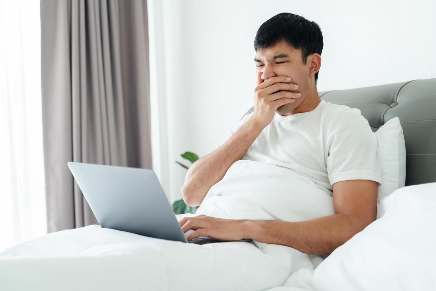 Sleepy man yawning on bed using laptop computer. photo